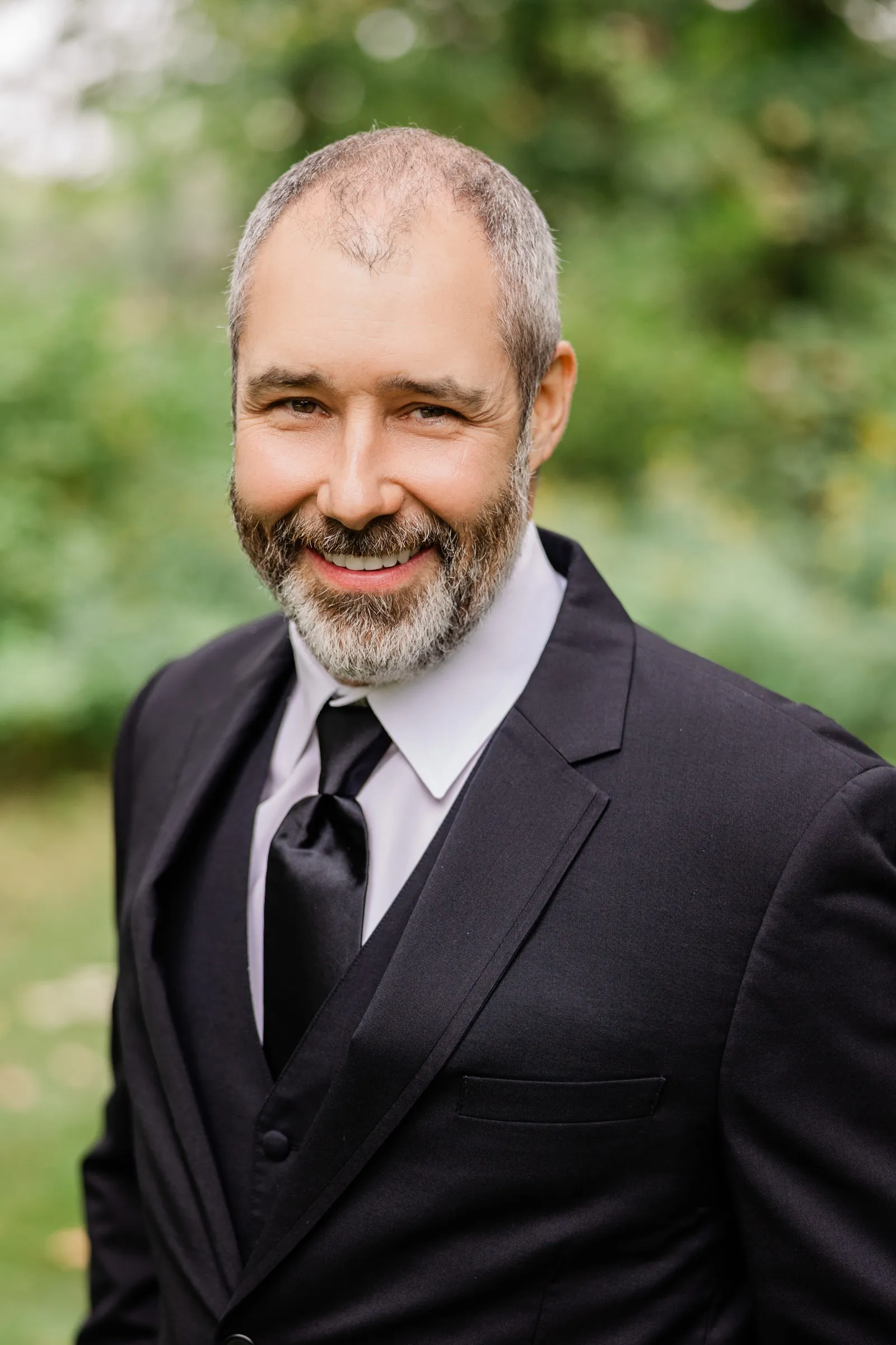 The groom in a black suit, vest and satin tie smiles at the camera against a soft green background. — Tim Larsen Photography, Brainerd Lakes MN