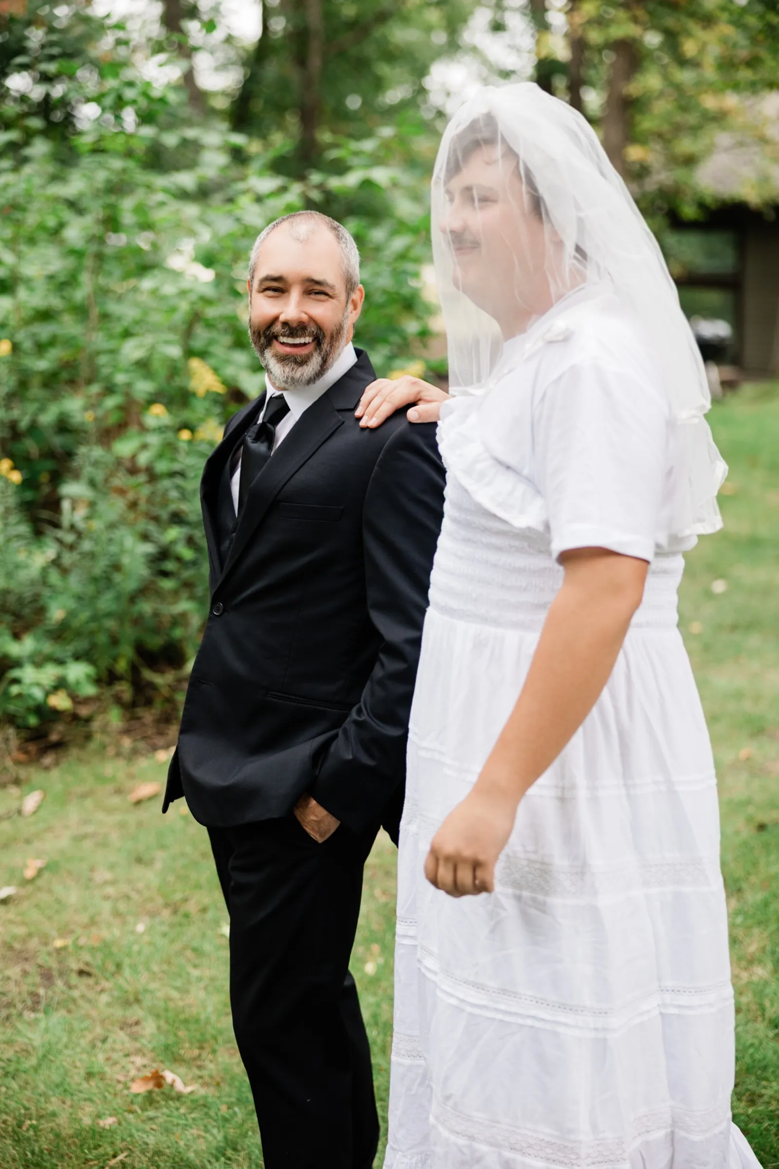 The groom laughs toward the camera as a young man with a mustache, wearing a white dress and a sheer veil, rests a hand on his shoulder. — Tim Larsen Photography, Brainerd Lakes MN