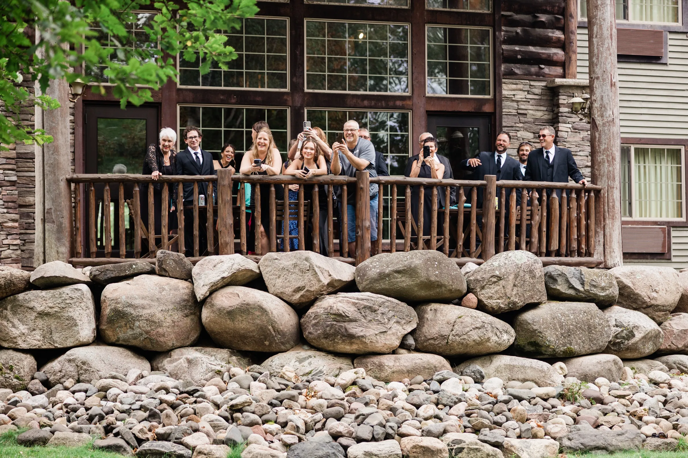 The wedding party and guests lean on a log railing on a deck above a boulder retaining wall, laughing and filming with phones in front of the lodge's tall windows. — Tim Larsen Photography, Brainerd Lakes MN