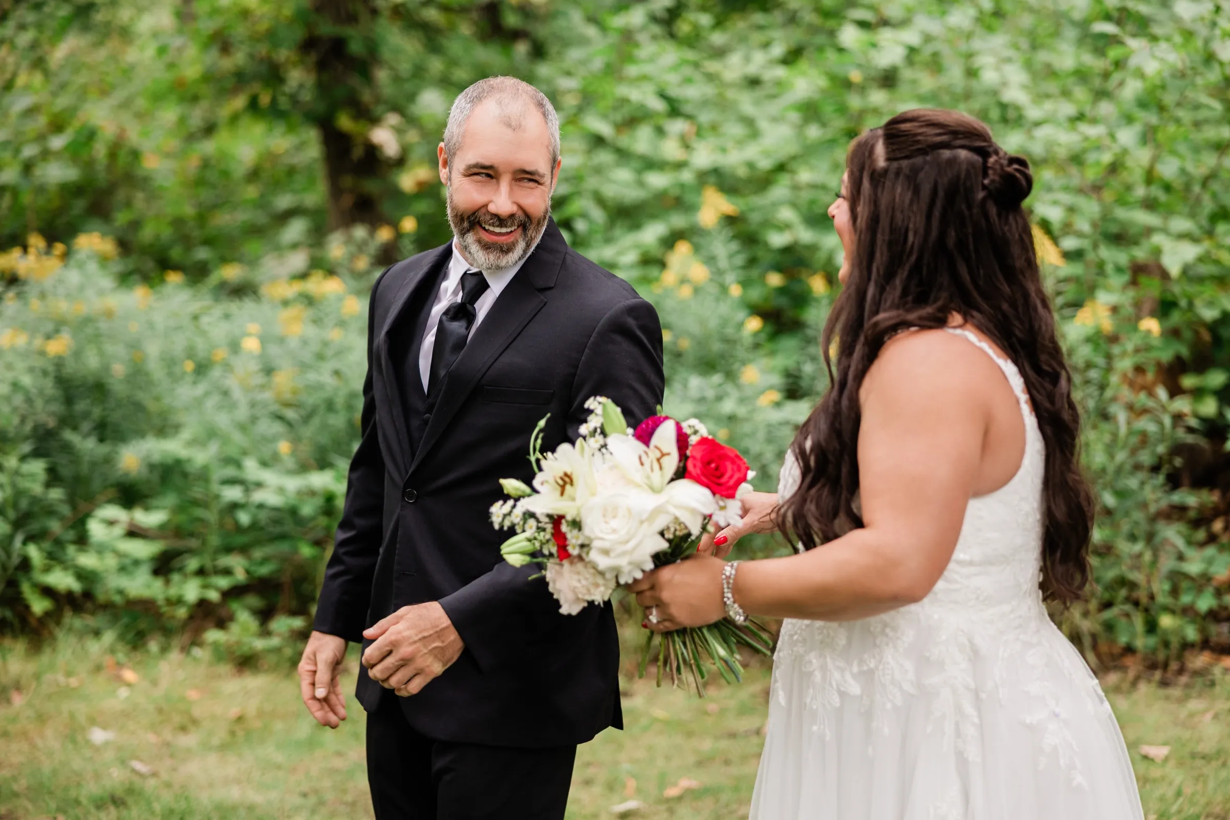 The groom turns and laughs broadly as he sees the bride for the first time, her long dark hair and bouquet in the foreground. — Tim Larsen Photography, Brainerd Lakes MN