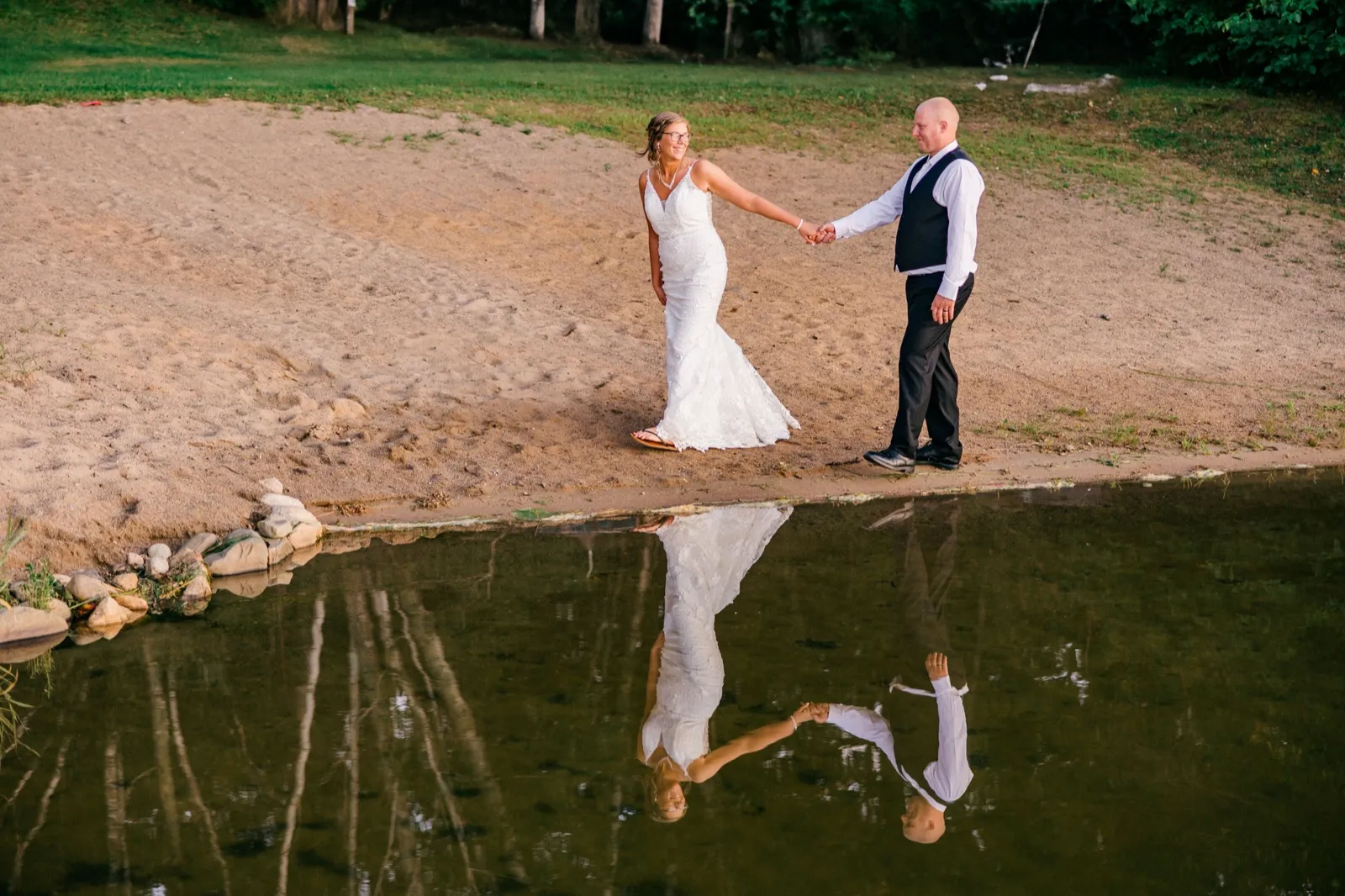 Kim and Jesse hold hands on a sandy lakeshore at golden hour, their full figures mirrored in the still water below — Tim Larsen Photography, Brainerd Lakes MN