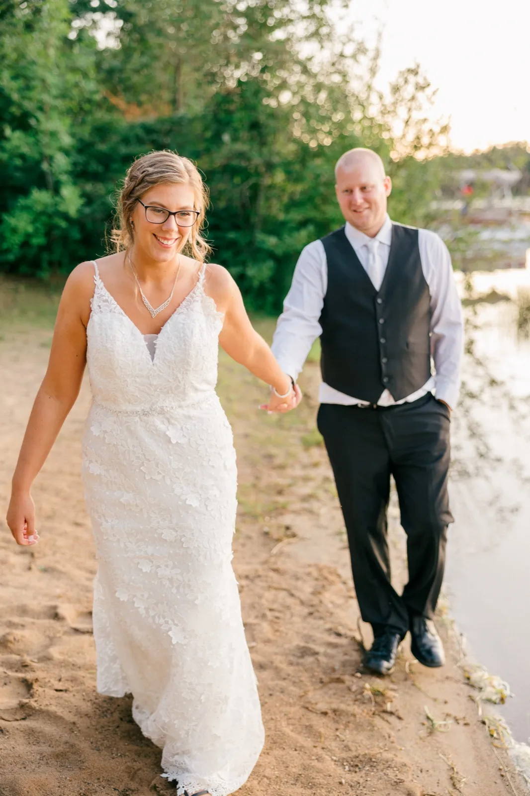 The bride leads the groom by the hand along a sandy lakeshore at golden hour, both smiling as warm light filters through the trees behind them — Tim Larsen Photography, Brainerd Lakes MN