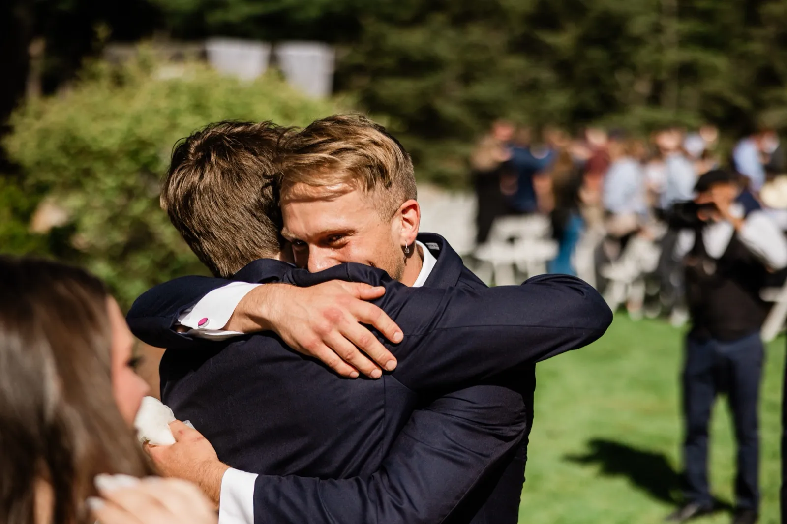 A groom holds a friend in a long, teary embrace on the ceremony lawn after the recessional, eyes shut — an unposed release caught while the crowd mingles behind them — Tim Larsen Photography, Brainerd Lakes MN