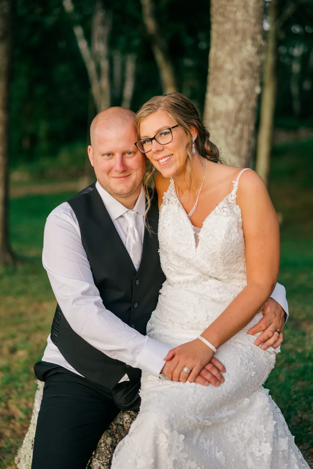 Kim leans her head against Jesse as they sit together on a log in a wooded grove, both smiling at the camera in soft evening light — Tim Larsen Photography, Brainerd Lakes MN