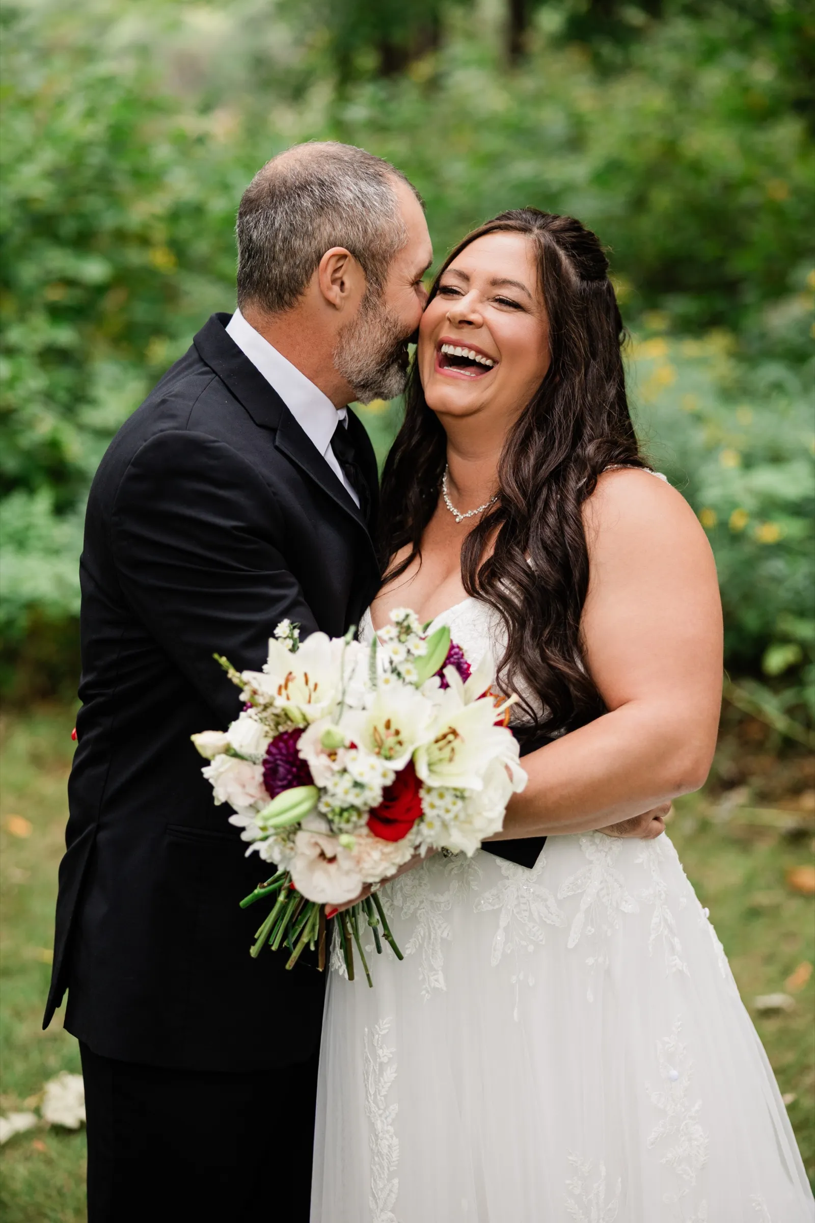 The bride throws her head back laughing while the groom kisses her cheek, her bouquet of white lilies, burgundy dahlias and red roses held in front of them. — Tim Larsen Photography, Brainerd Lakes MN