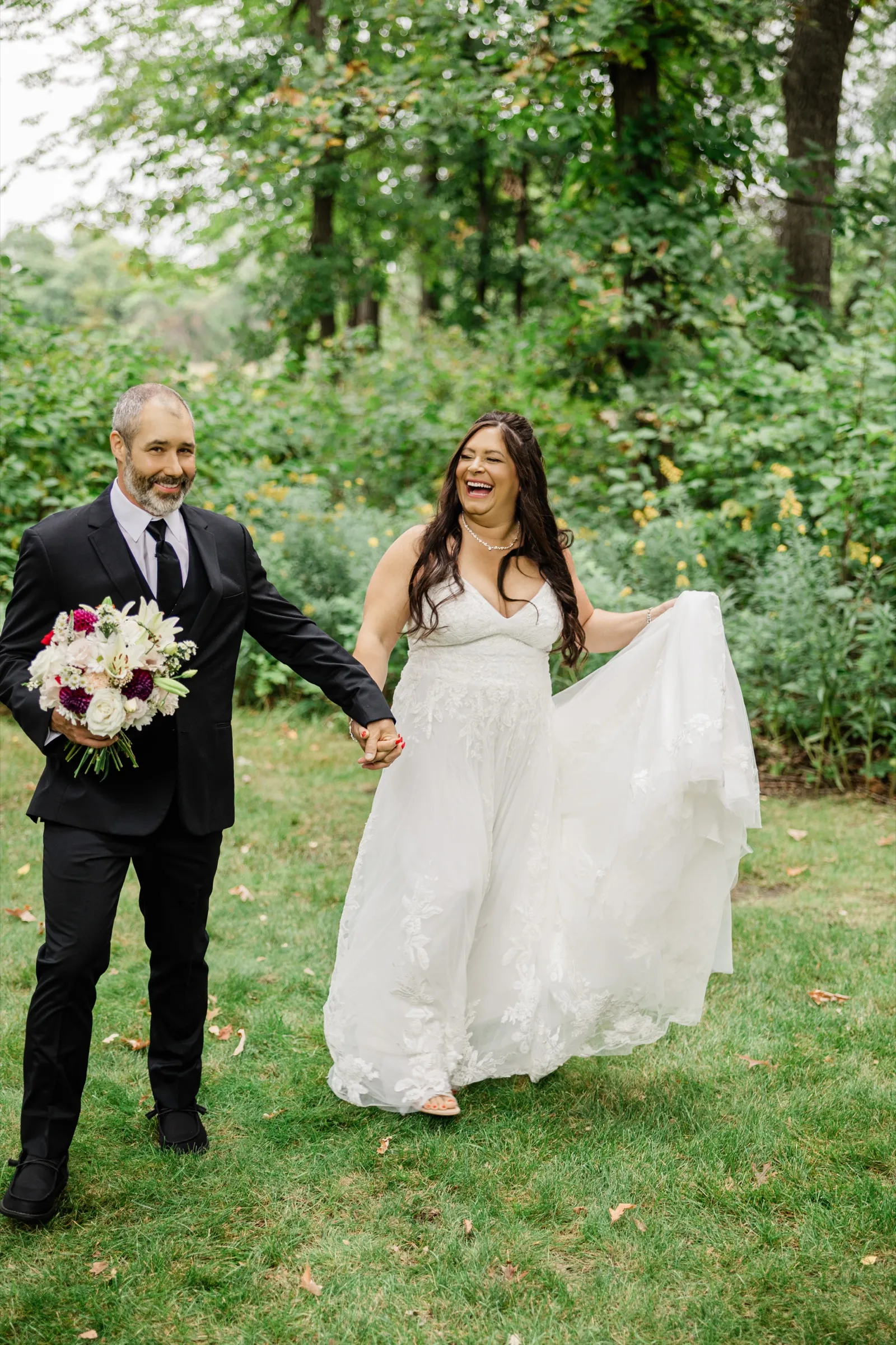 The groom leads the bride by the hand across the lawn, smiling at the camera, while she laughs openly and lifts her skirt with her other hand. — Tim Larsen Photography, Brainerd Lakes MN