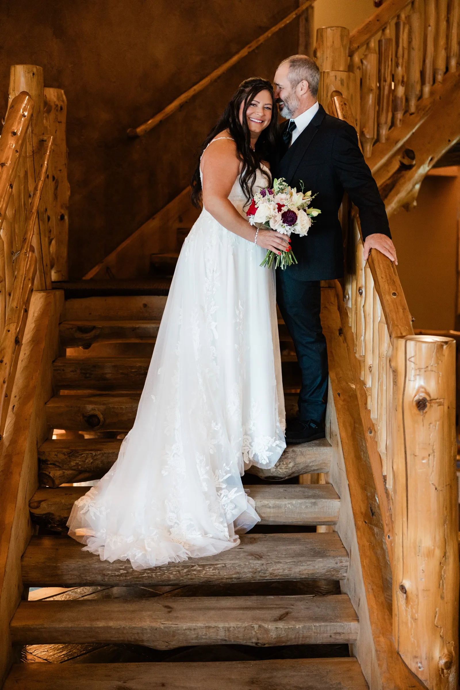 The bride smiles at the camera while the groom leans in close to her on a staircase of thick wooden treads and hand-peeled log railings inside the lodge. — Tim Larsen Photography, Brainerd Lakes MN