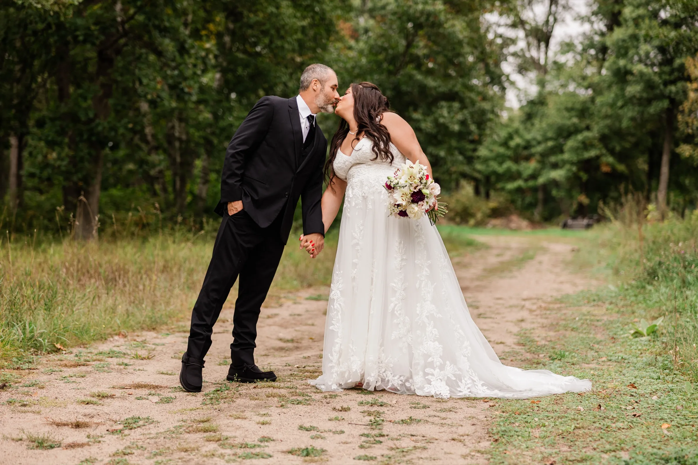 Holding hands, the couple leans in for a kiss on a sandy dirt path that curves into a stand of tall trees. — Tim Larsen Photography, Brainerd Lakes MN
