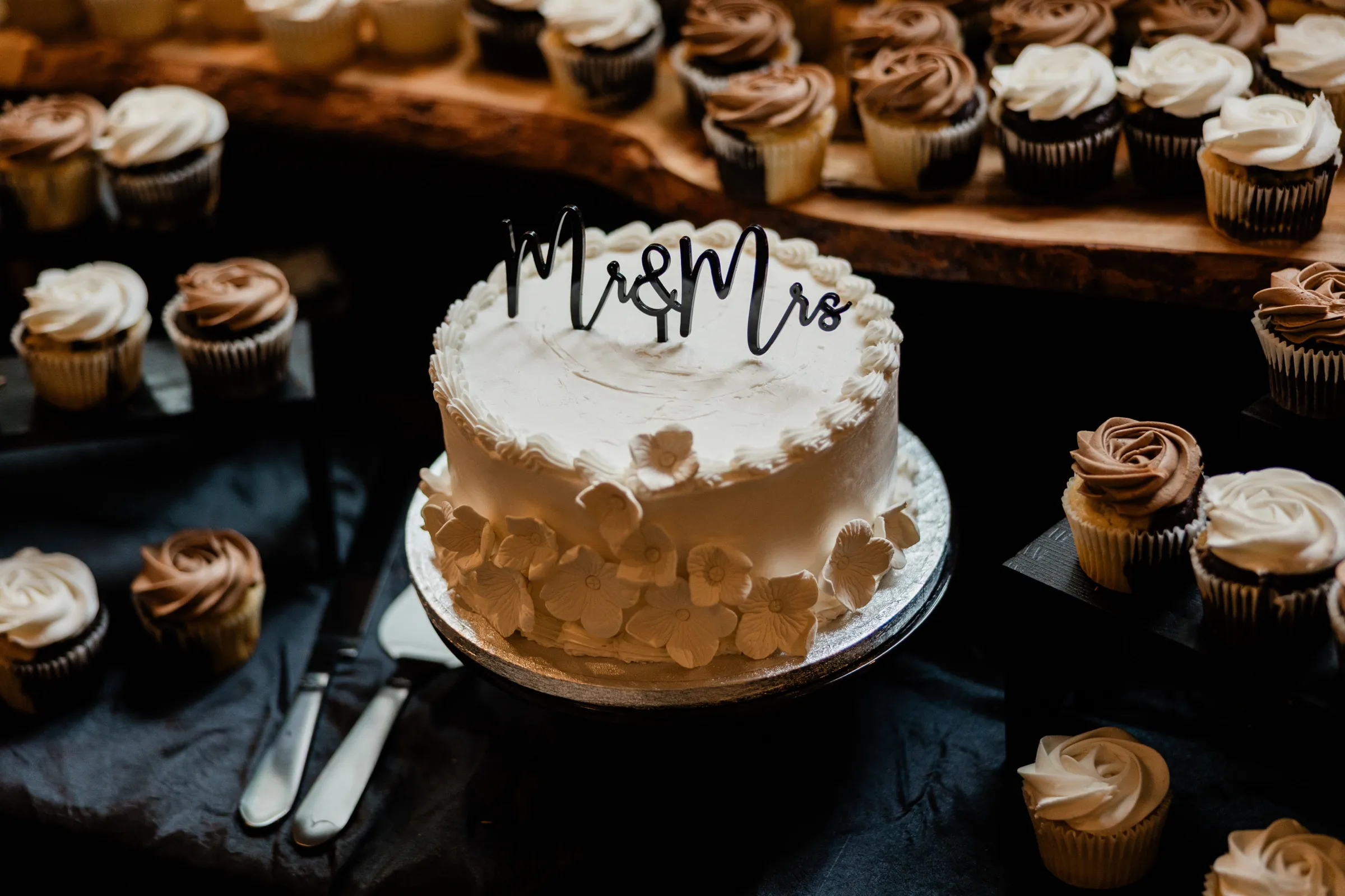 A single-tier white cake with sugar flowers and a black script Mr & Mrs topper sits on a silver board, surrounded by chocolate and vanilla cupcakes on wooden slabs and black cloth. — Tim Larsen Photography, Brainerd Lakes MN