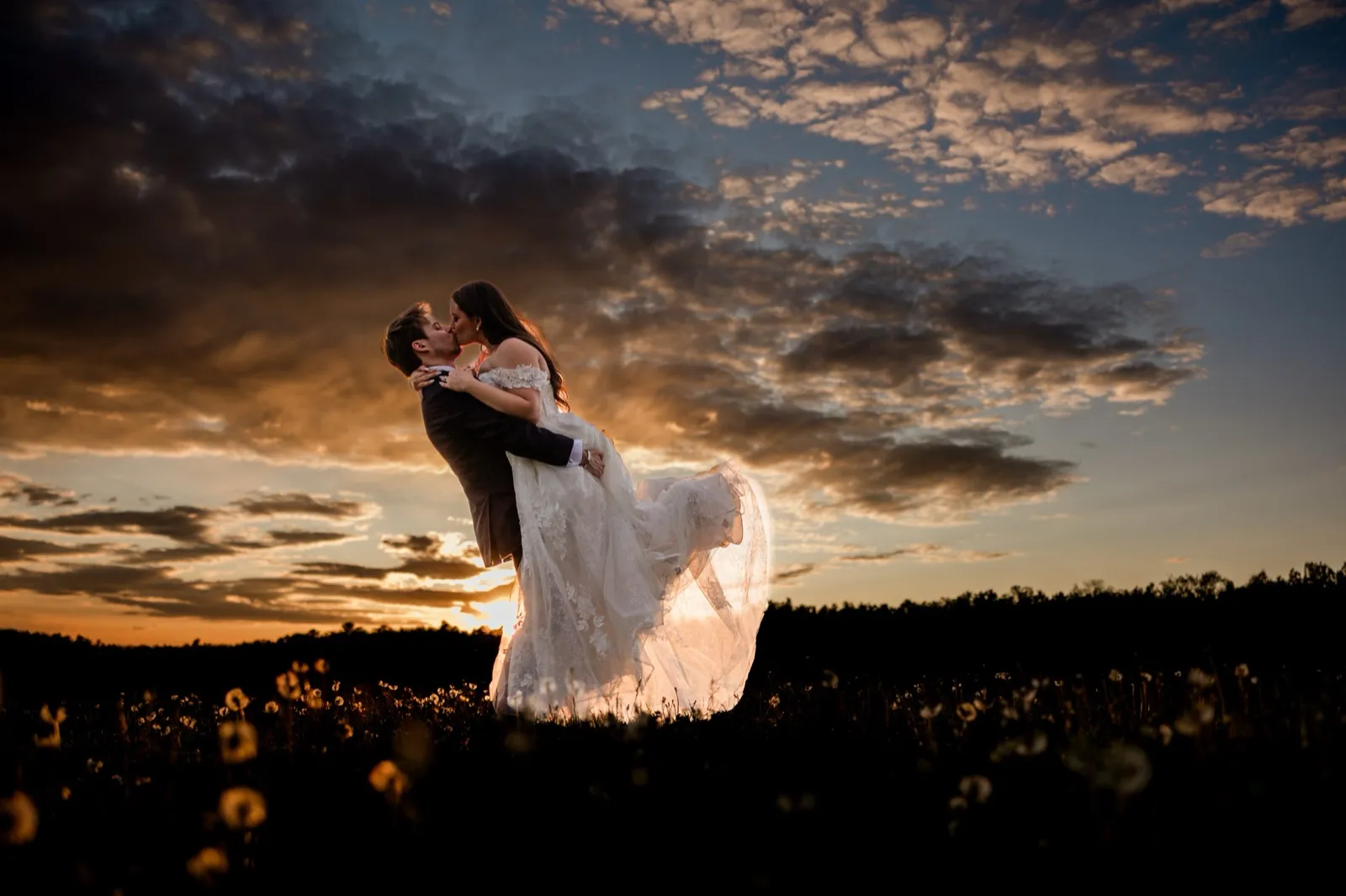 A groom lifts the bride in a wildflower field as the sun sets behind them, the sky streaked with cloud and gold — the longer, richer golden hour fall gives back — Tim Larsen Photography, Brainerd Lakes MN