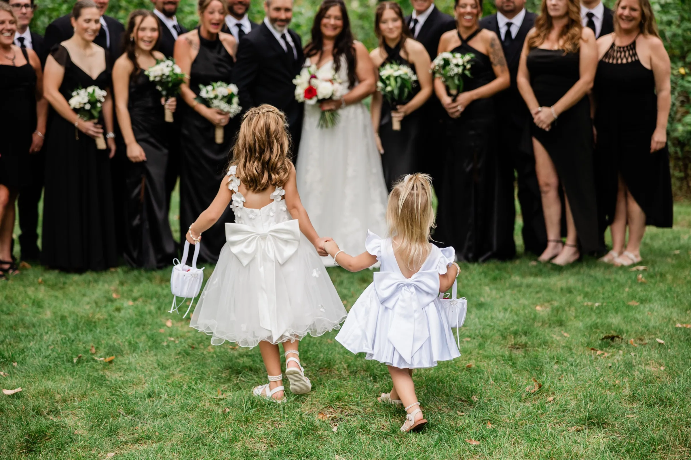 Seen from behind, two small flower girls with large white bows on their dresses walk hand in hand across the grass toward the couple and the smiling wedding party. — Tim Larsen Photography, Brainerd Lakes MN
