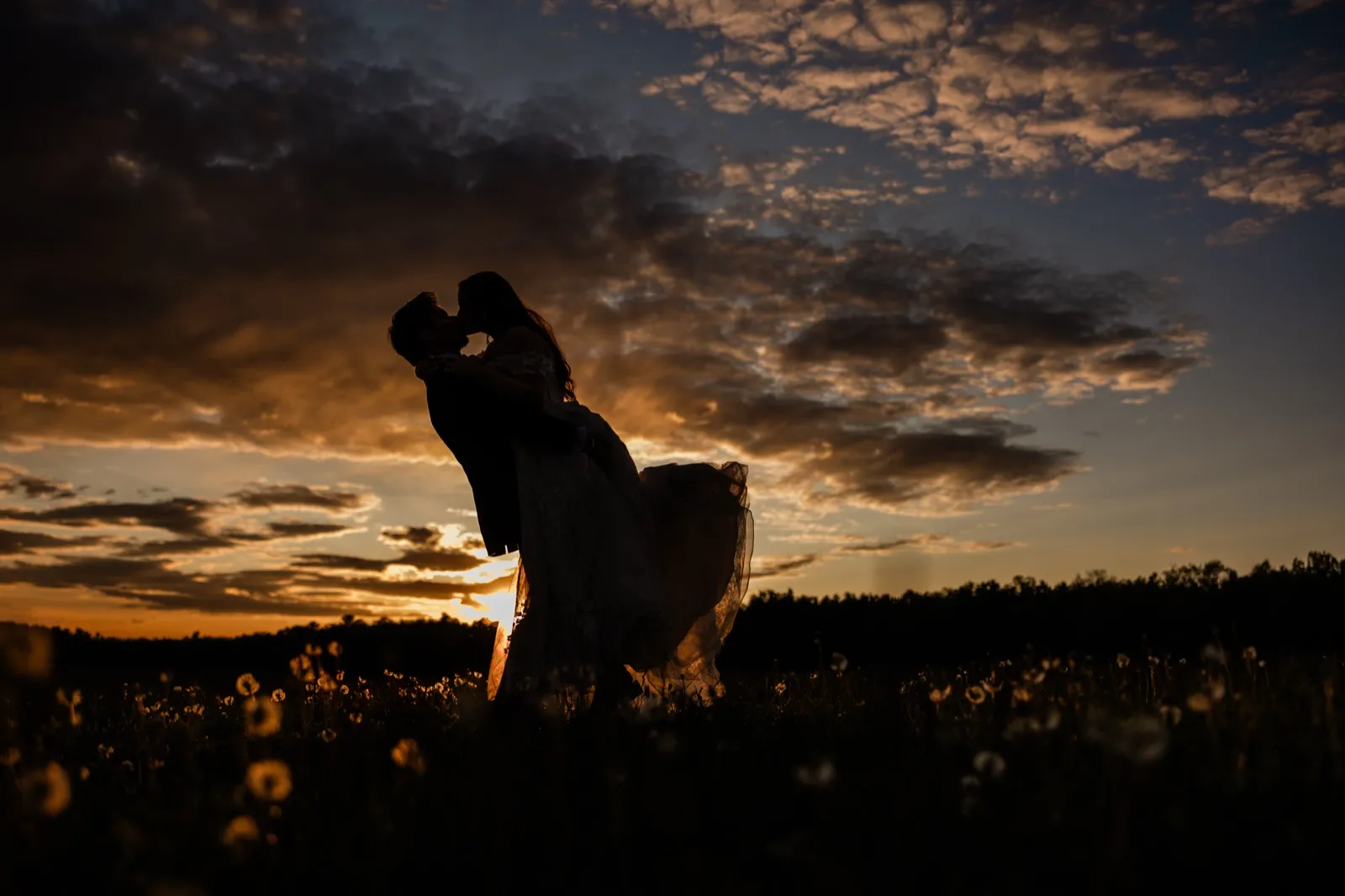 Silhouettes of the couple kissing against a blazing orange sunset sky over the Pine Peaks meadow