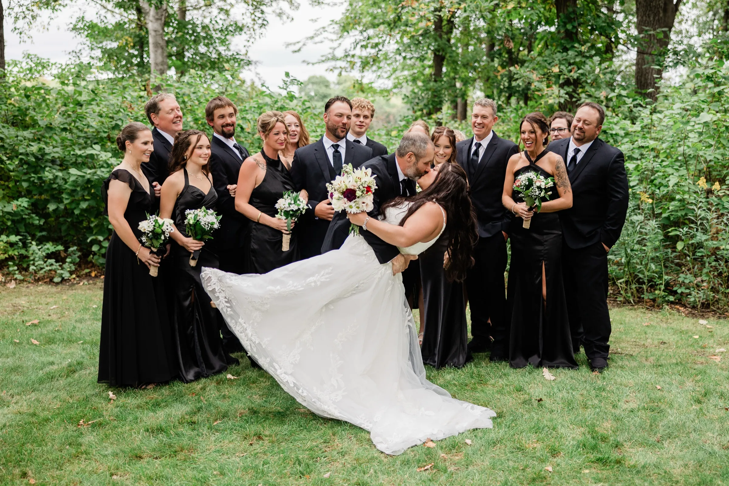 The groom dips the bride into a kiss on the lawn, her lace dress sweeping out across the grass, while the wedding party behind them laughs and cheers. — Tim Larsen Photography, Brainerd Lakes MN