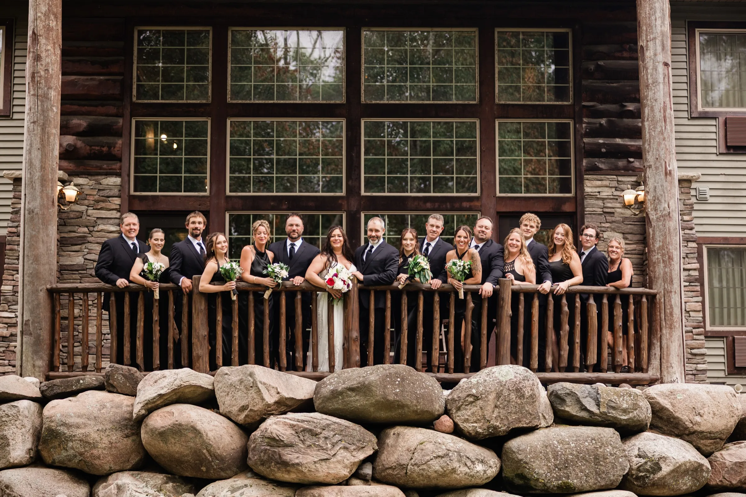 The couple and their wedding party lean along a log railing on the lodge deck, tall grid windows and log-and-stone walls behind them and a boulder wall below. — Tim Larsen Photography, Brainerd Lakes MN