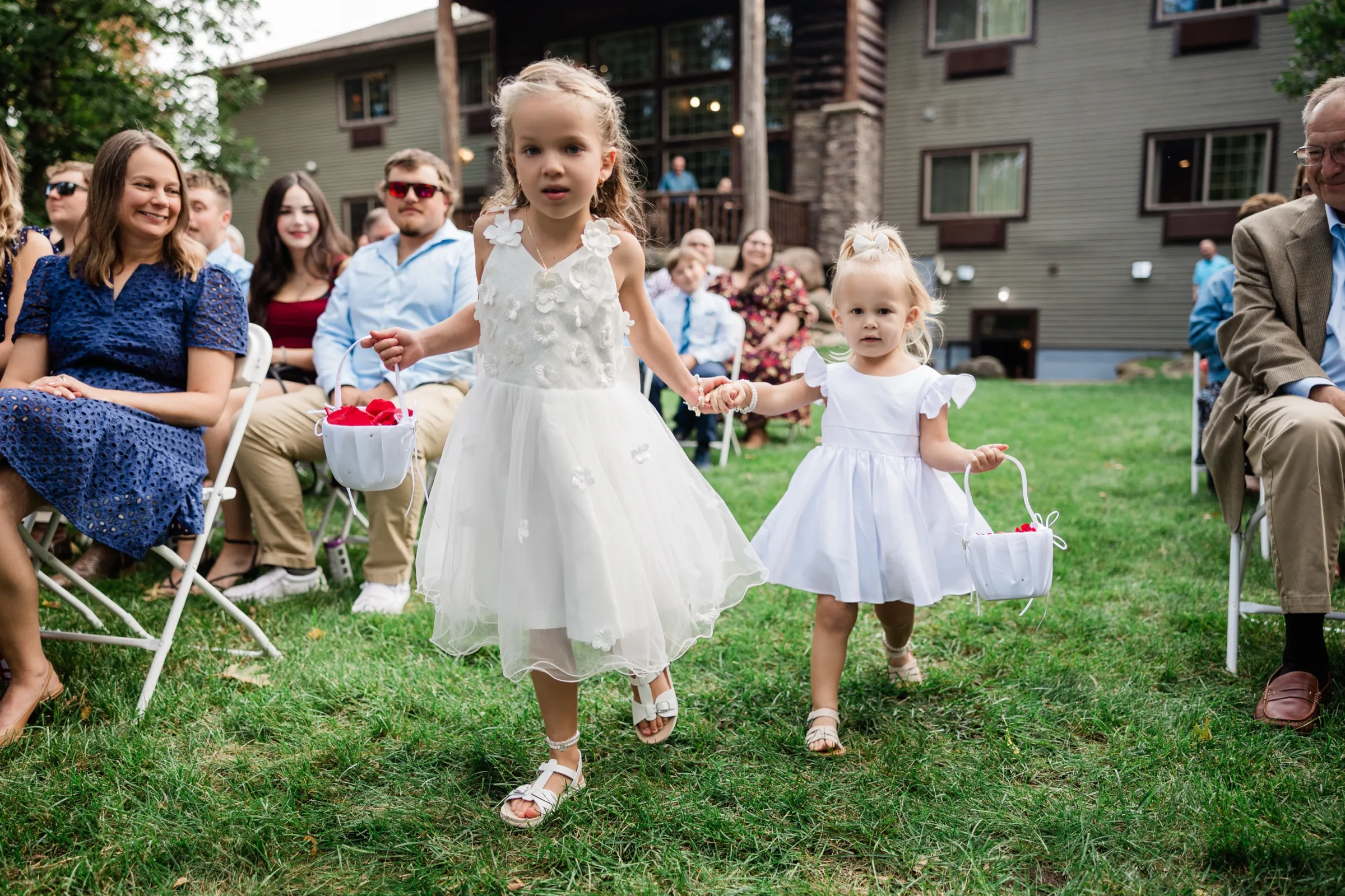 Two young flower girls in white dresses hold hands and carry petal baskets down the lawn aisle as seated guests smile, with the lodge building behind them. — Tim Larsen Photography, Brainerd Lakes MN
