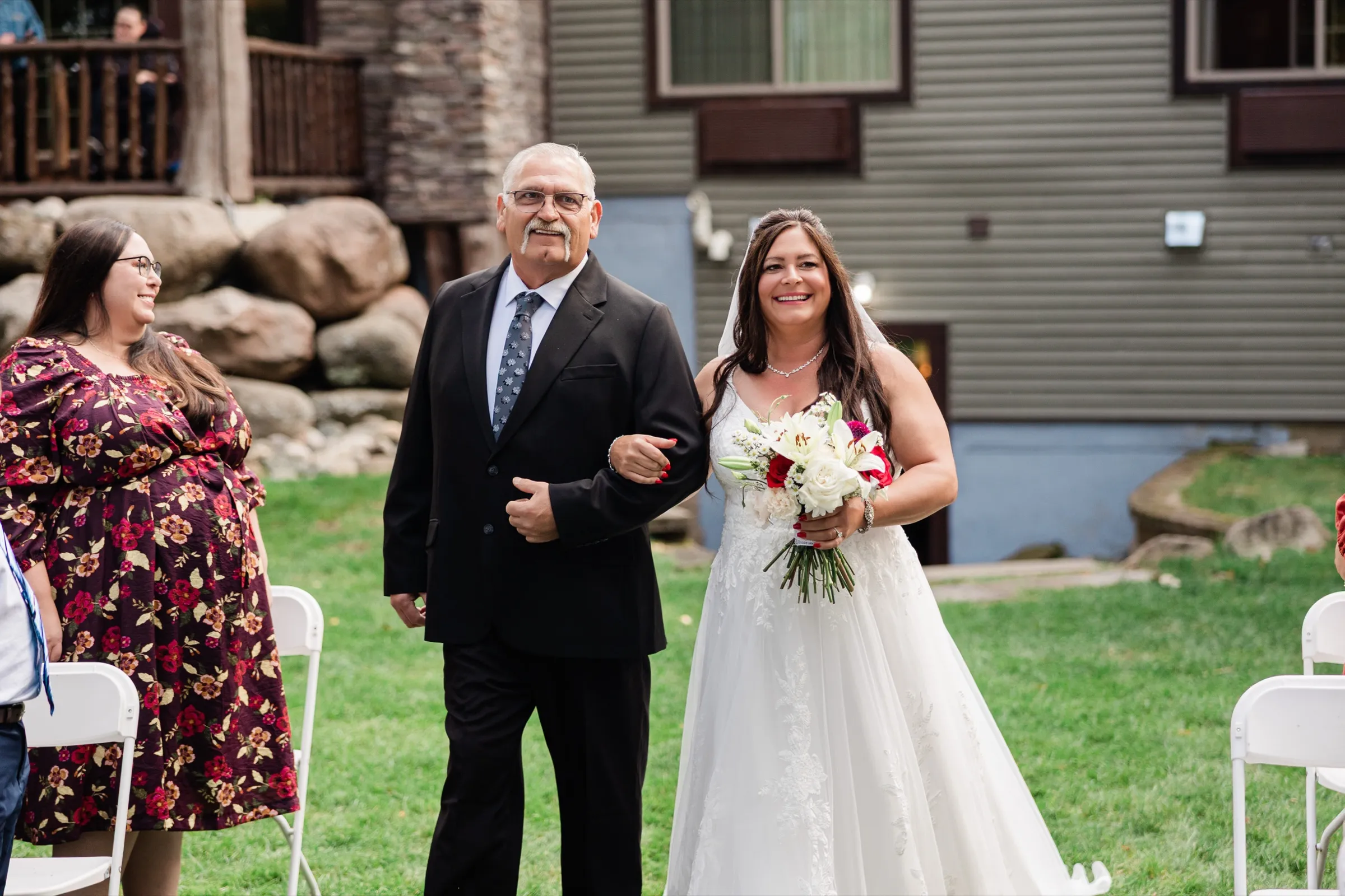 Amber smiles as she walks down the lawn aisle on the arm of an older man in a dark suit, holding a bouquet of white lilies and red roses. — Tim Larsen Photography, Brainerd Lakes MN