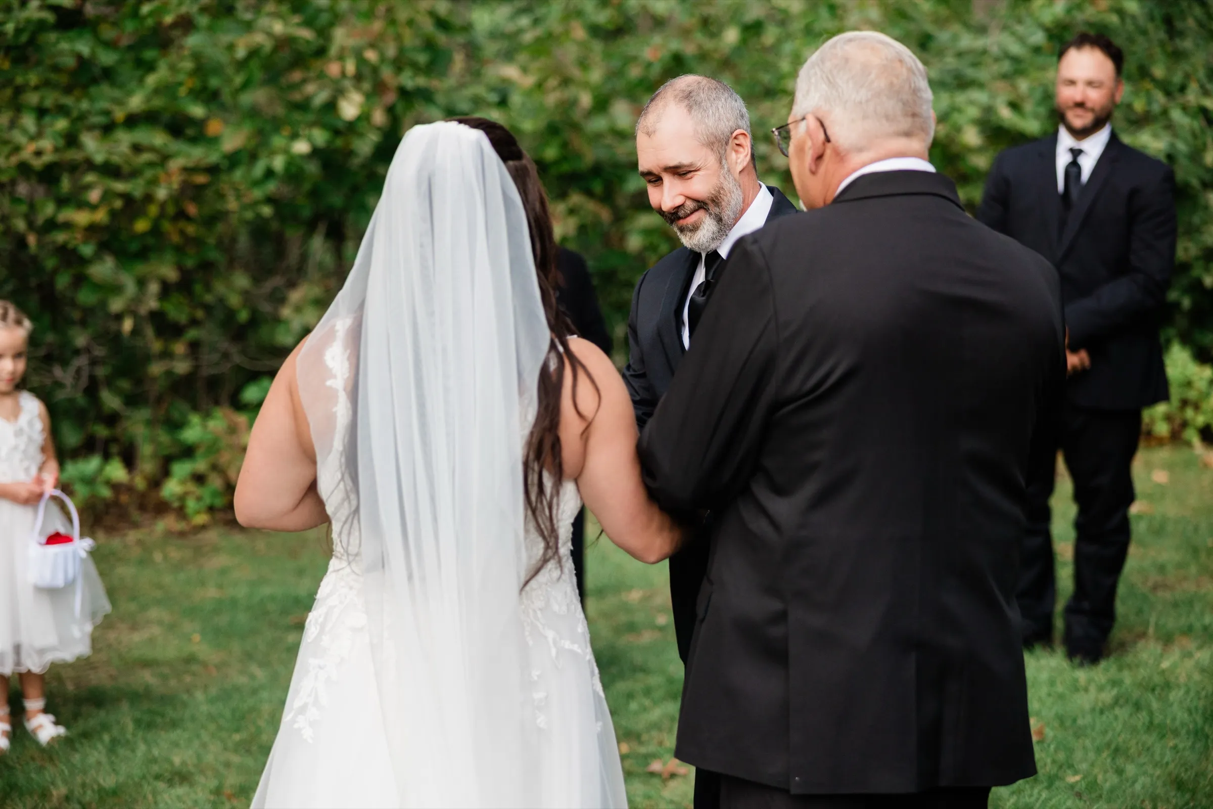 Don smiles and looks down as he reaches for Amber at the front of the ceremony, seen past her veil and the shoulder of the man who walked her in. — Tim Larsen Photography, Brainerd Lakes MN