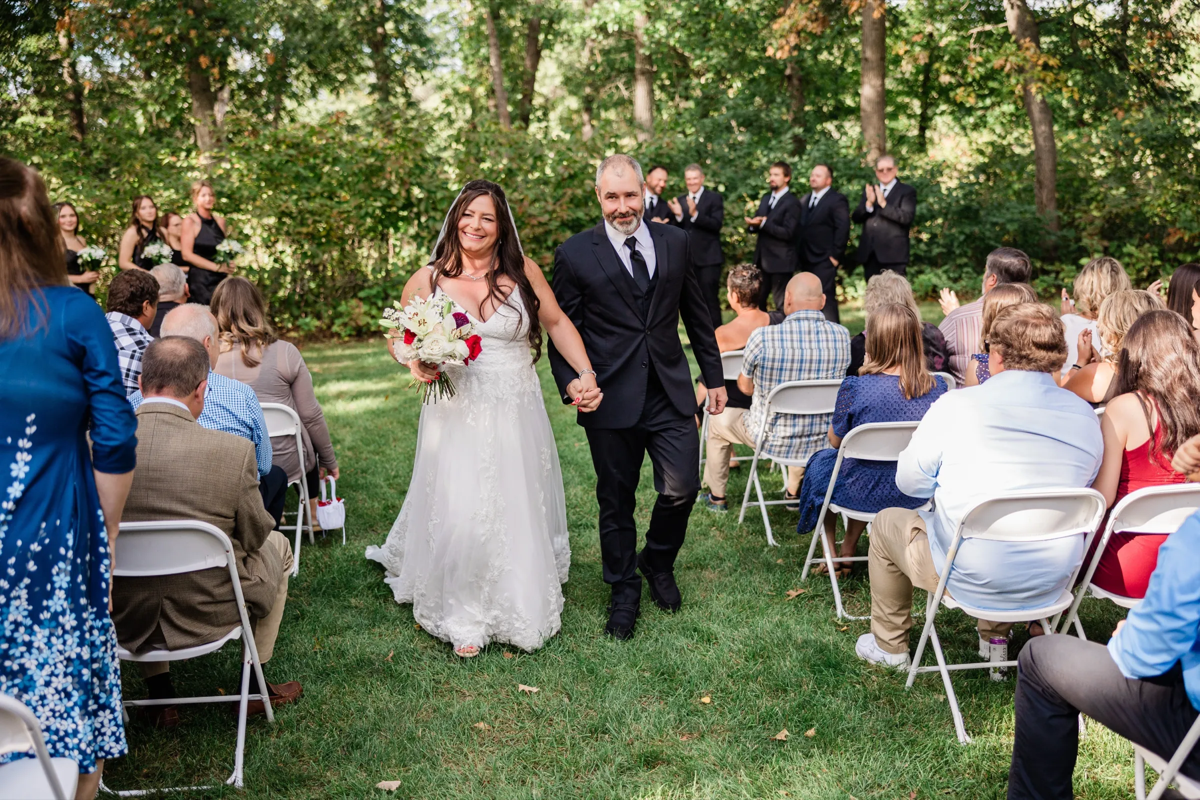 Amber and Don walk hand in hand back up the lawn aisle between rows of guests on white chairs as the bridal party applauds behind them. — Tim Larsen Photography, Brainerd Lakes MN