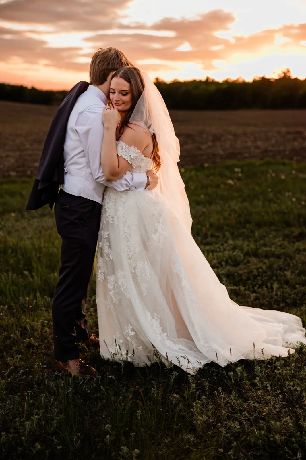 Bride resting her head on the groom's chest in an open field beneath a vivid sunset sky