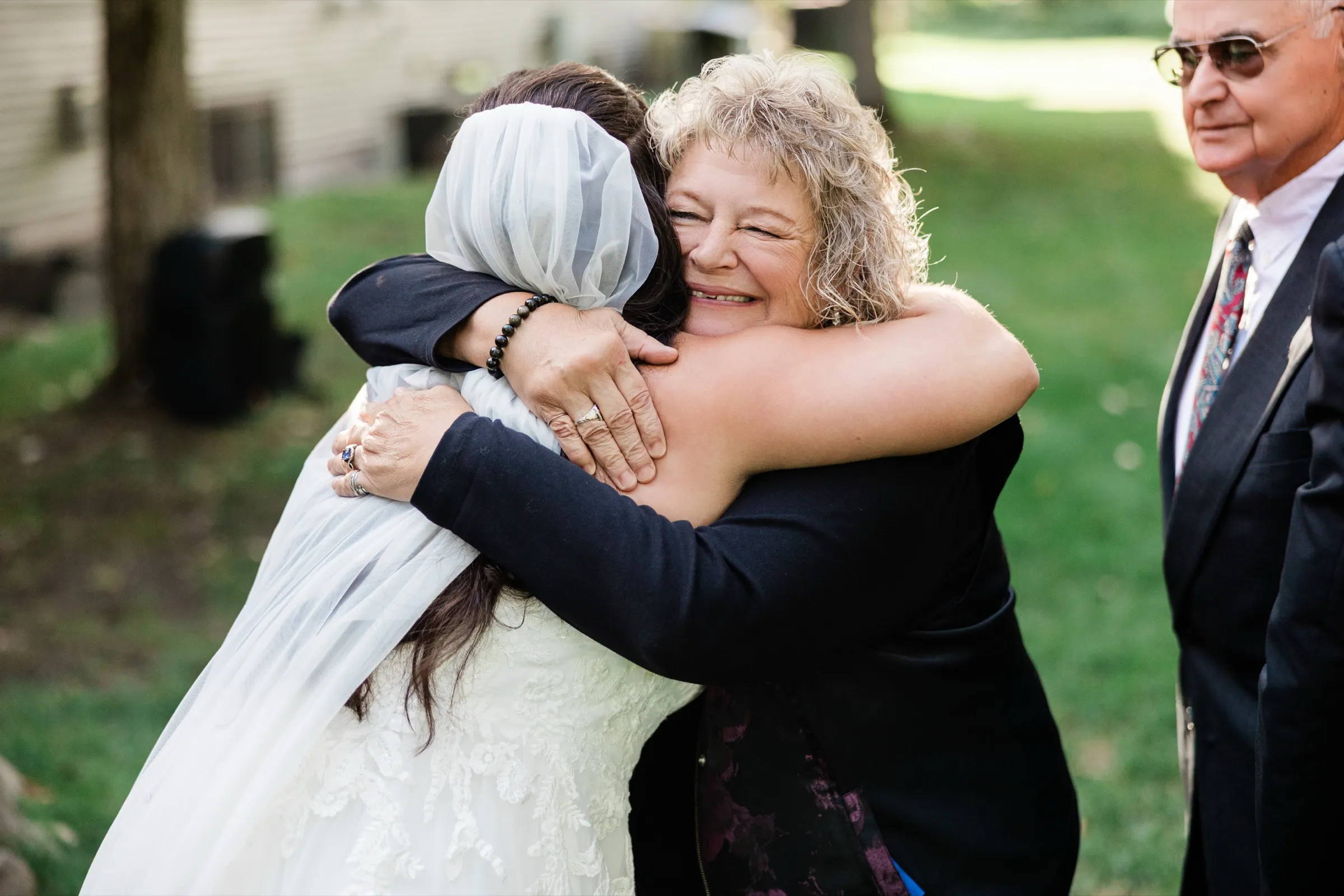 A woman with curly blonde hair squeezes her eyes shut and grins as she hugs Amber, while an older man in a suit looks on. — Tim Larsen Photography, Brainerd Lakes MN