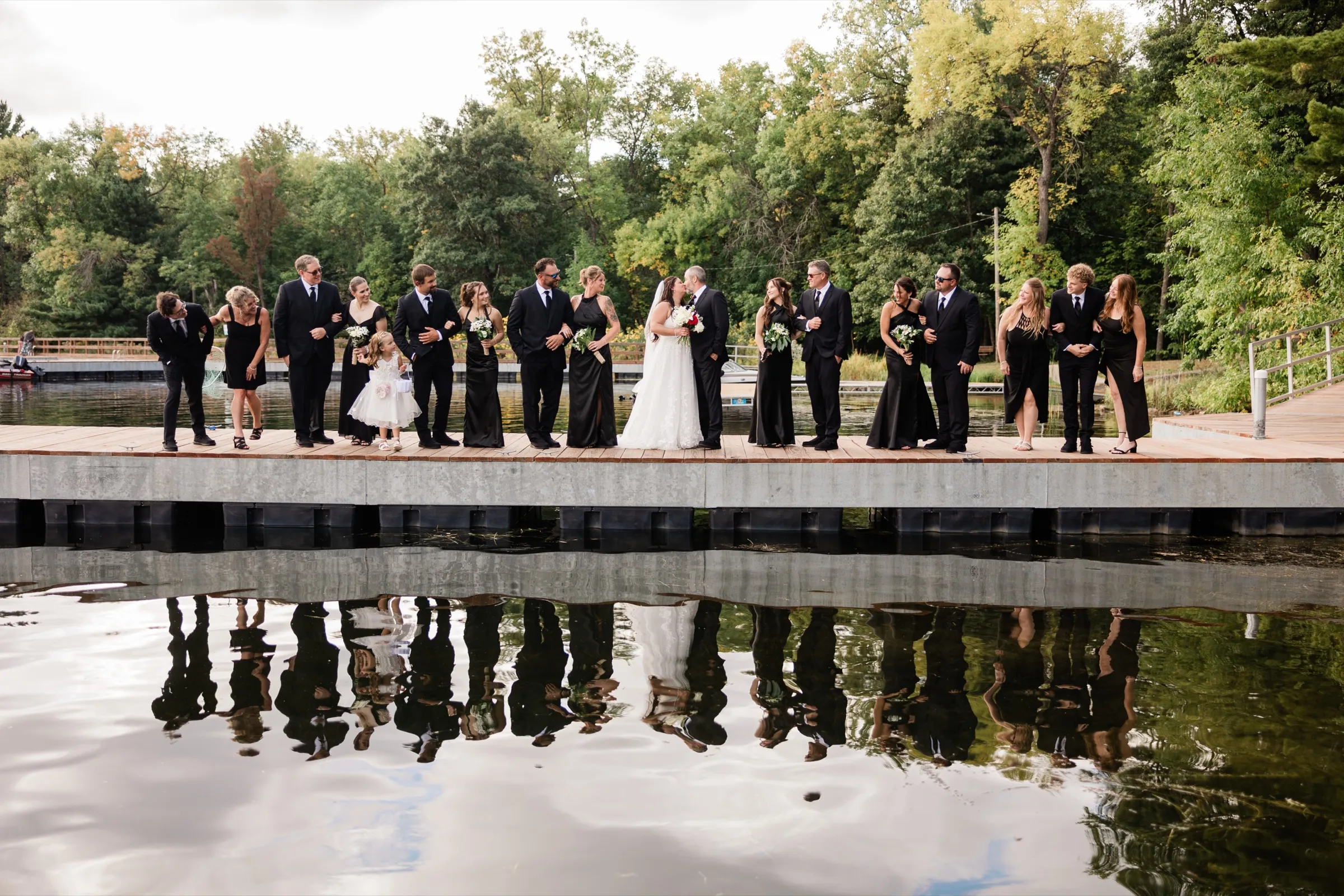 The full bridal party stands arm in arm along a wooden dock, reflected in the still lake below, while Amber and Don kiss at the center. — Tim Larsen Photography, Brainerd Lakes MN