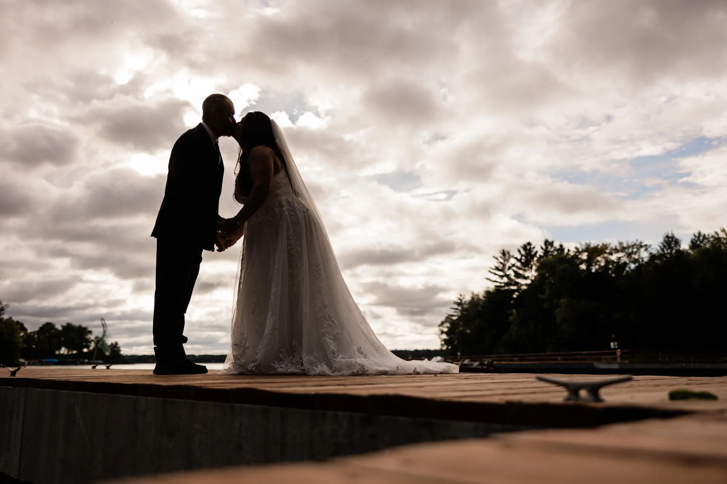 Amber and Don kiss in silhouette at the end of a dock under a sky full of heavy clouds, with a metal cleat in the foreground. — Tim Larsen Photography, Brainerd Lakes MN