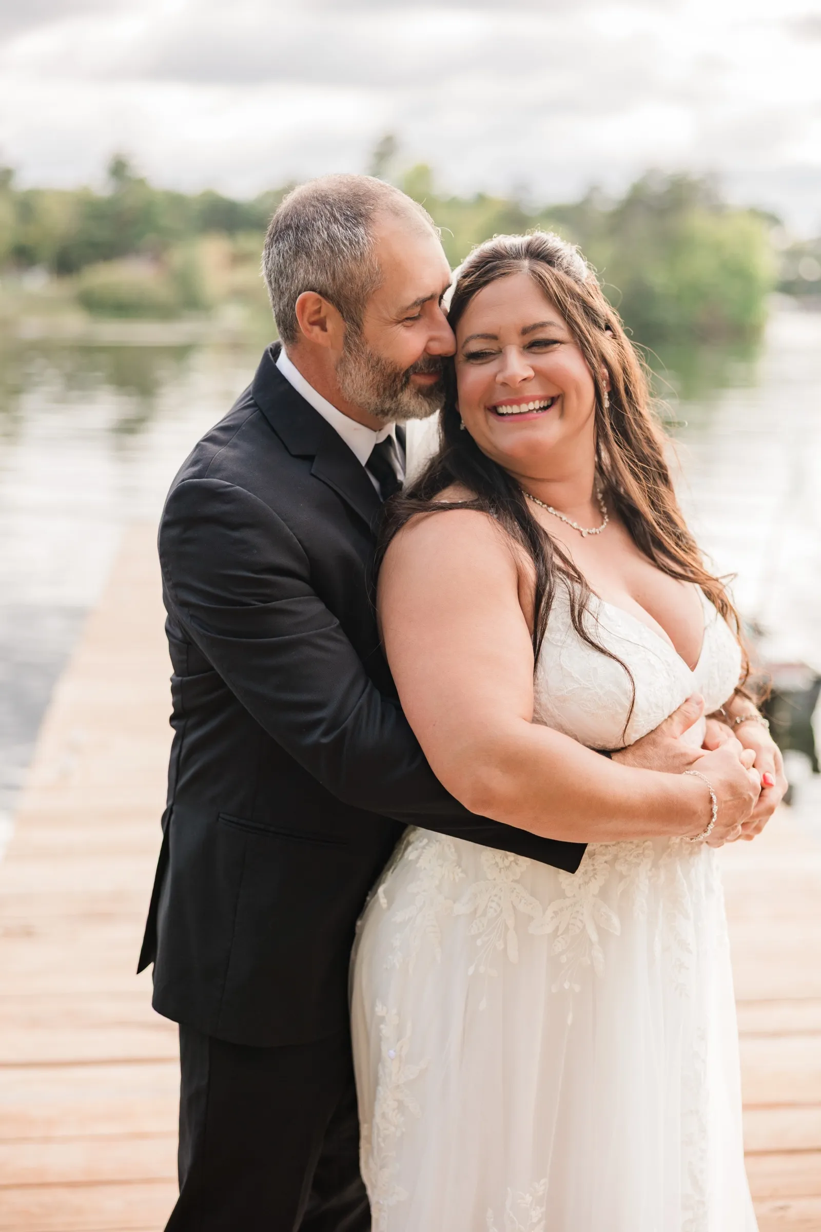 Don wraps his arms around Amber from behind and presses his face to hers as she laughs on the dock, with the lake softly blurred behind them. — Tim Larsen Photography, Brainerd Lakes MN