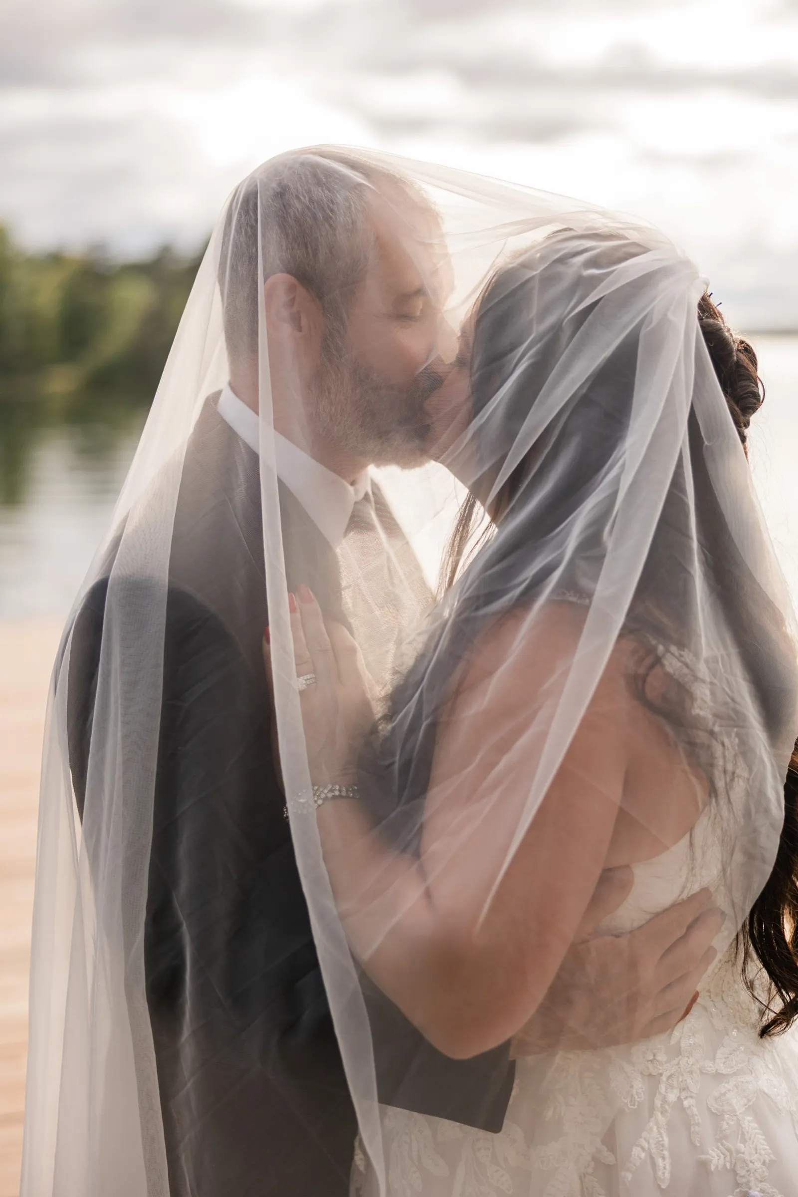 Amber and Don kiss beneath her sheer veil, which is draped over both of them and lit from behind by the cloudy sky over the lake. — Tim Larsen Photography, Brainerd Lakes MN