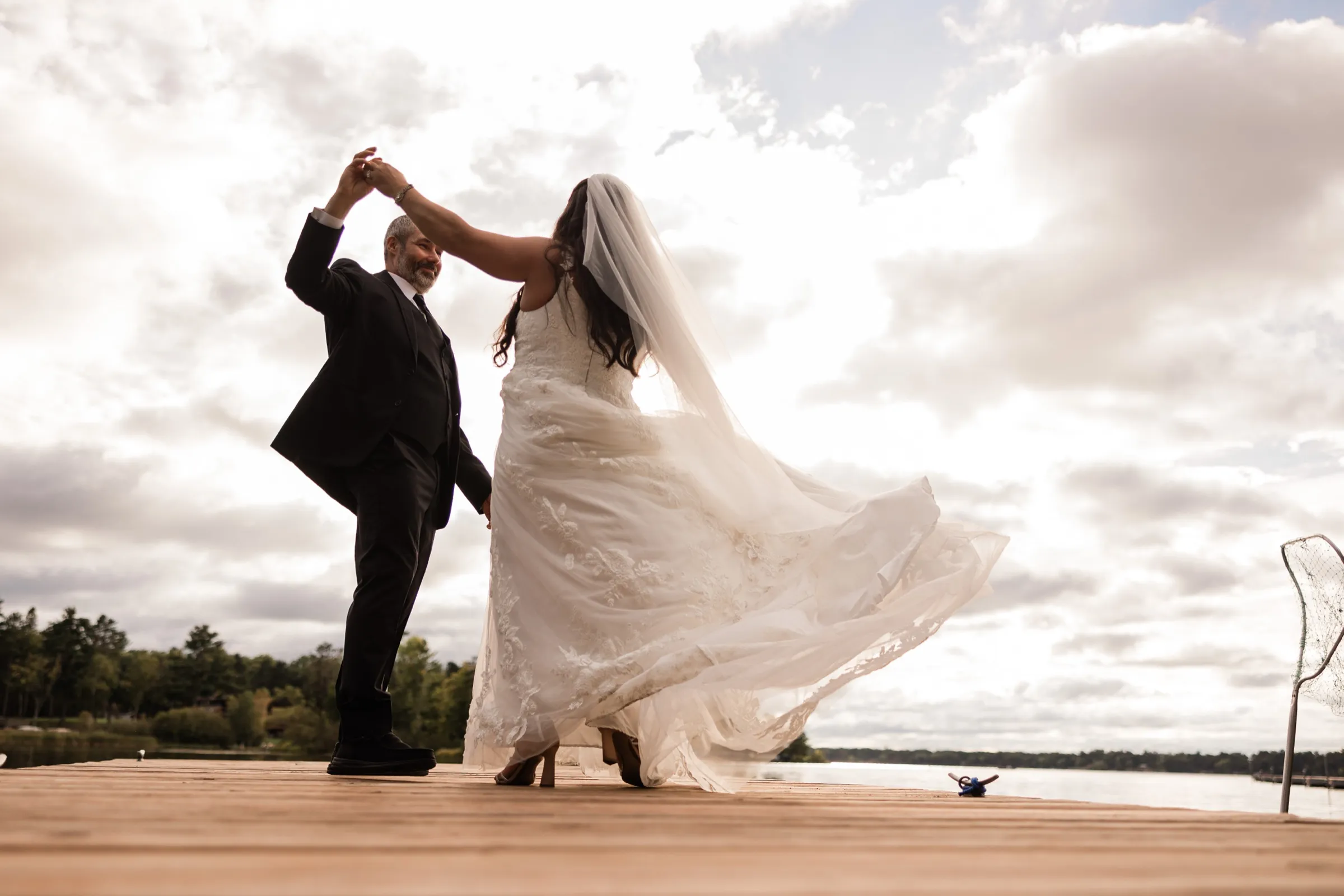 Don twirls Amber at the end of a dock, her lace dress and long veil flaring out against a sky of dramatic clouds over the lake. — Tim Larsen Photography, Brainerd Lakes MN