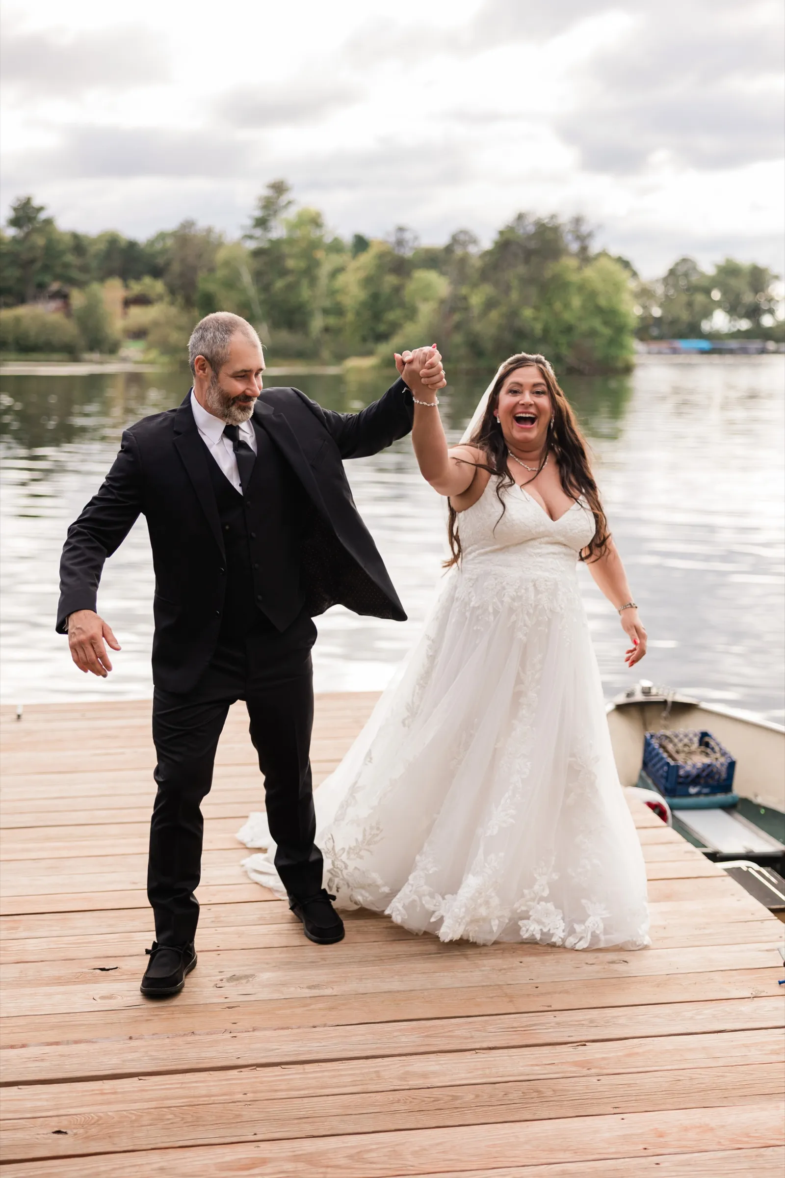 Amber cheers with her mouth wide open as she and Don raise their joined hands on a wooden dock beside a small boat, with the lake behind them. — Tim Larsen Photography, Brainerd Lakes MN
