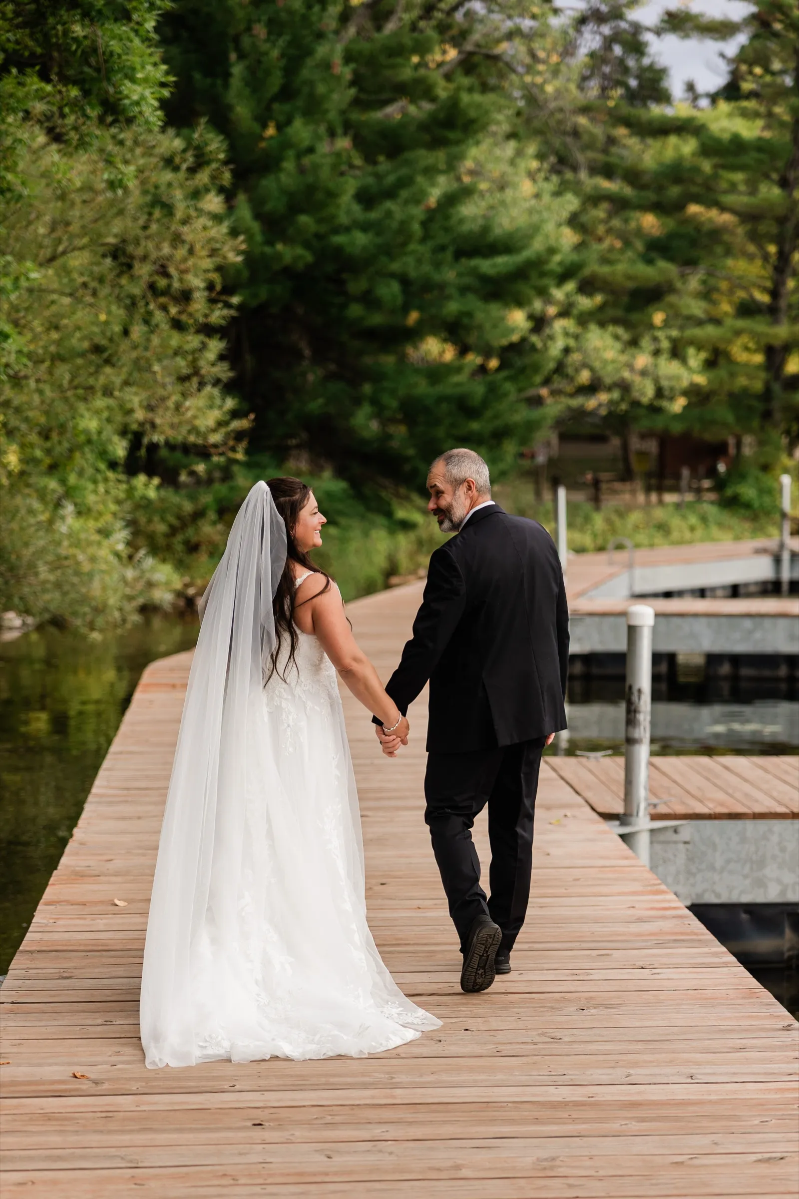 Amber and Don look back at each other and smile as they walk hand in hand along a wooden dock beneath tall pines. — Tim Larsen Photography, Brainerd Lakes MN