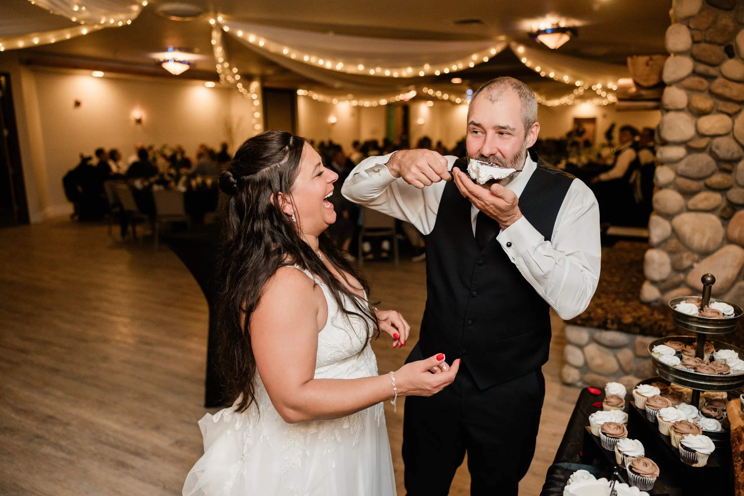 Don takes an oversized bite of cake from a fork while Amber laughs with her head tipped back beside the cupcake table. — Tim Larsen Photography, Brainerd Lakes MN