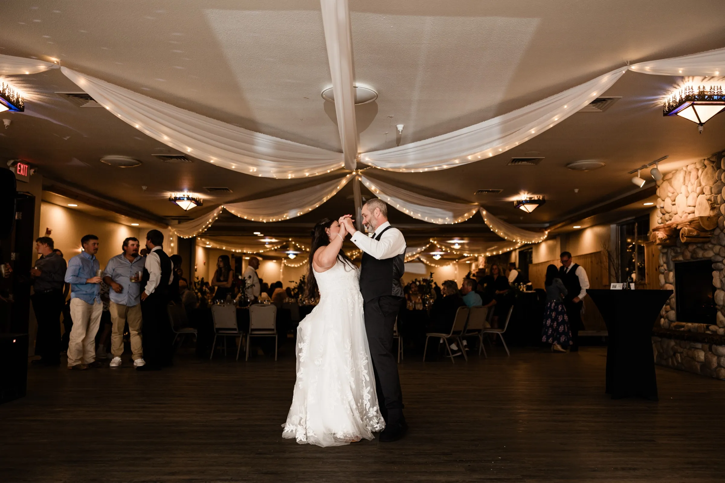 Amber and Don dance alone on the floor of a ballroom with draped white fabric and string lights on the ceiling and a stone fireplace on the right, guests watching from the edges. — Tim Larsen Photography, Brainerd Lakes MN
