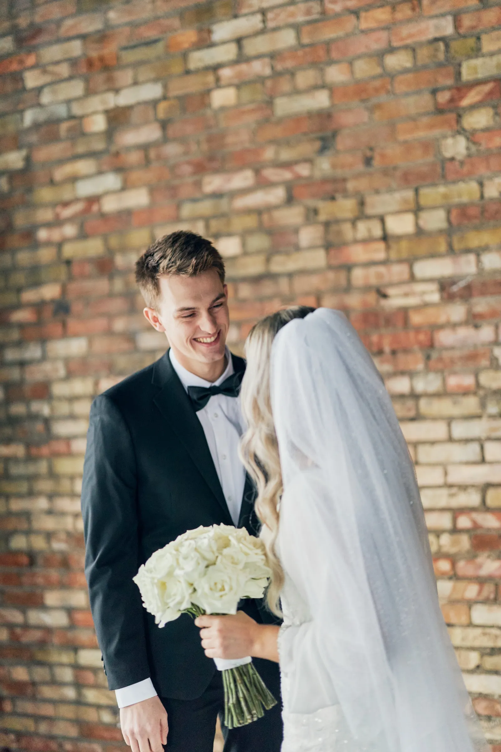 Sam smiles warmly at Madeline, who faces him in a long veil holding a white rose bouquet against a weathered brick wall — Brainerd Lakes wedding portraits — Tim Larsen Photography, Brainerd Lakes MN