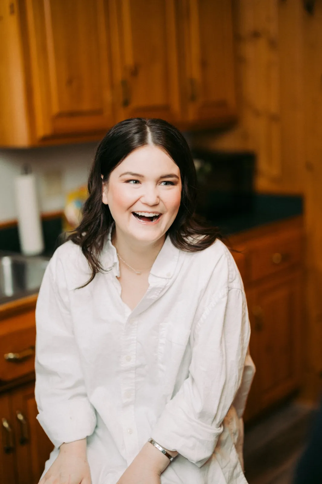 The bride laughs openly while getting ready in a warmly lit pine kitchen, wearing an oversized white button-down shirt — Tim Larsen Photography, Brainerd Lakes MN