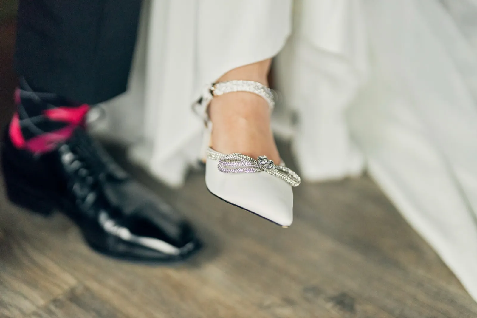 A close-up of the bride's jeweled white heel lifted beside the groom's black patent oxford as they stand close together on a wood floor. — Tim Larsen Photography, Brainerd Lakes MN