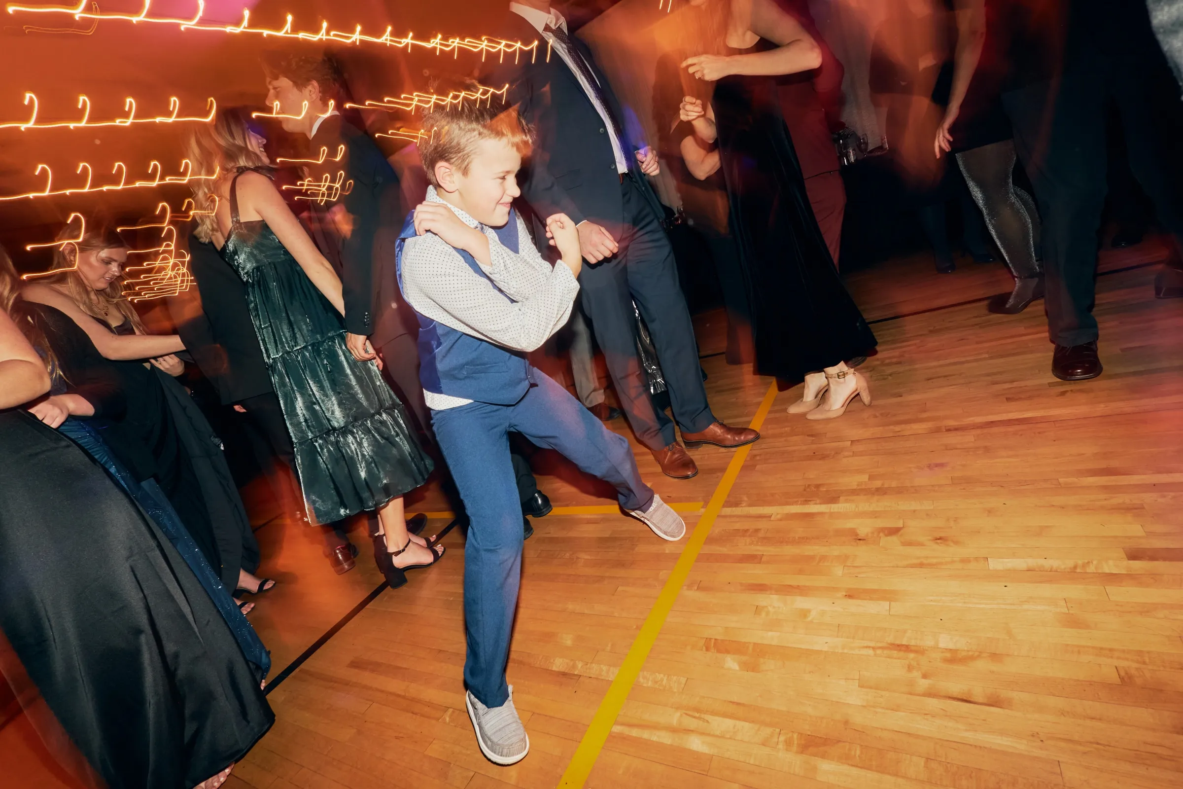 A young boy in a vest and dress pants strikes a confident dance pose on the wooden gym floor as guests dance around him under streaking string lights — Tim Larsen Photography, Brainerd Lakes MN