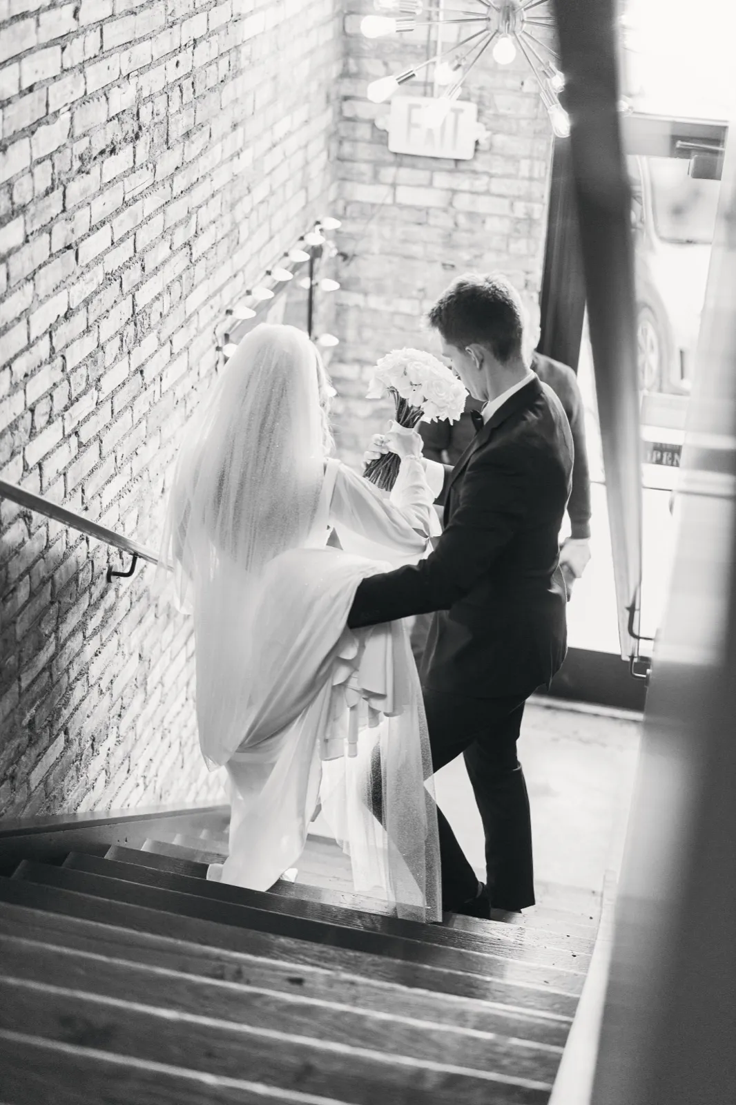 A black-and-white image looking down a staircase as the bride and groom descend together, her veil and white bouquet trailing across the steps beneath an exposed brick wall and sputnik chandelier. — Tim Larsen Photography, Brainerd Lakes MN