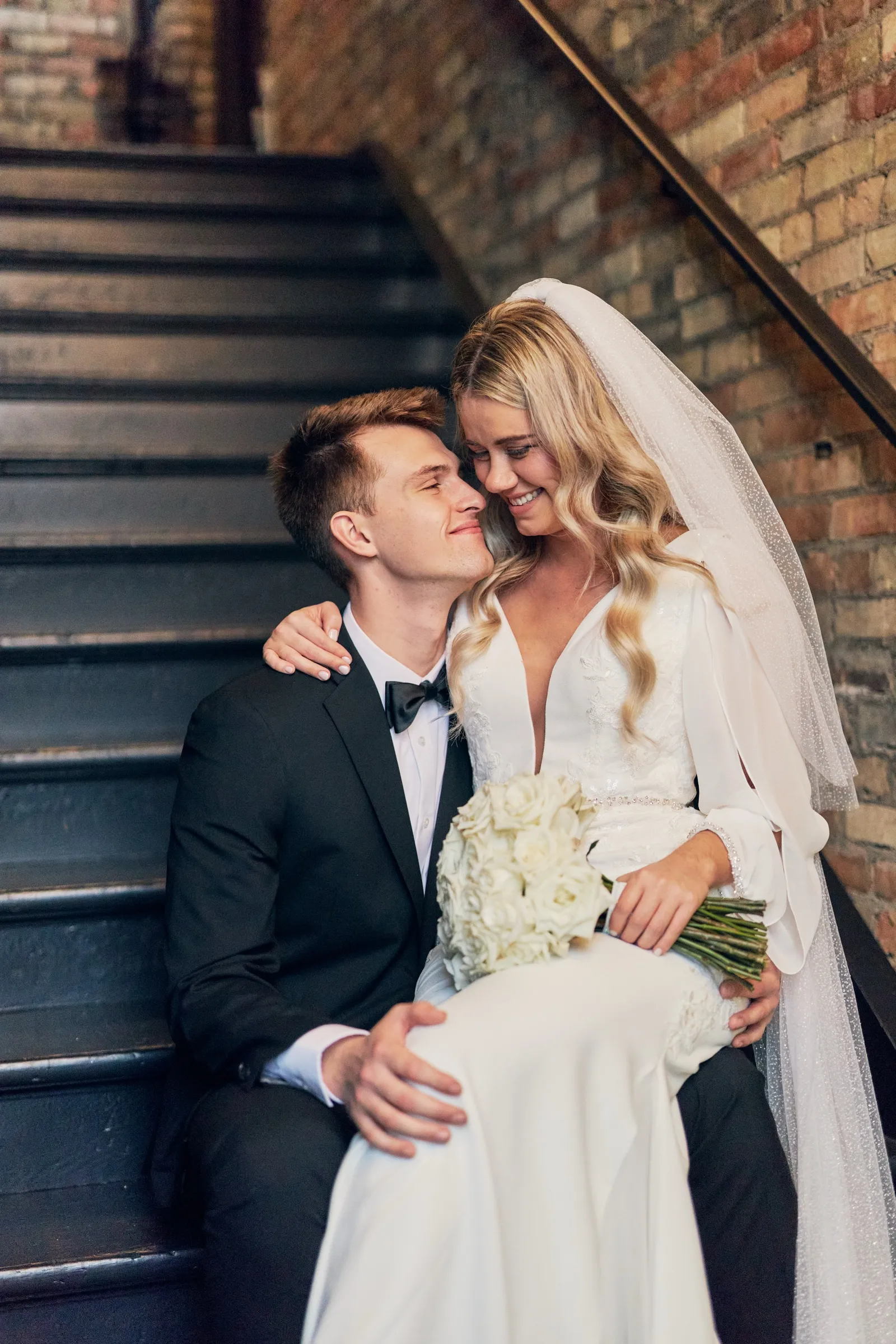 Madeline and Sam sit close together on a dark wooden staircase against an exposed brick wall, her veil flowing down the steps — Tim Larsen Photography, Brainerd Lakes MN