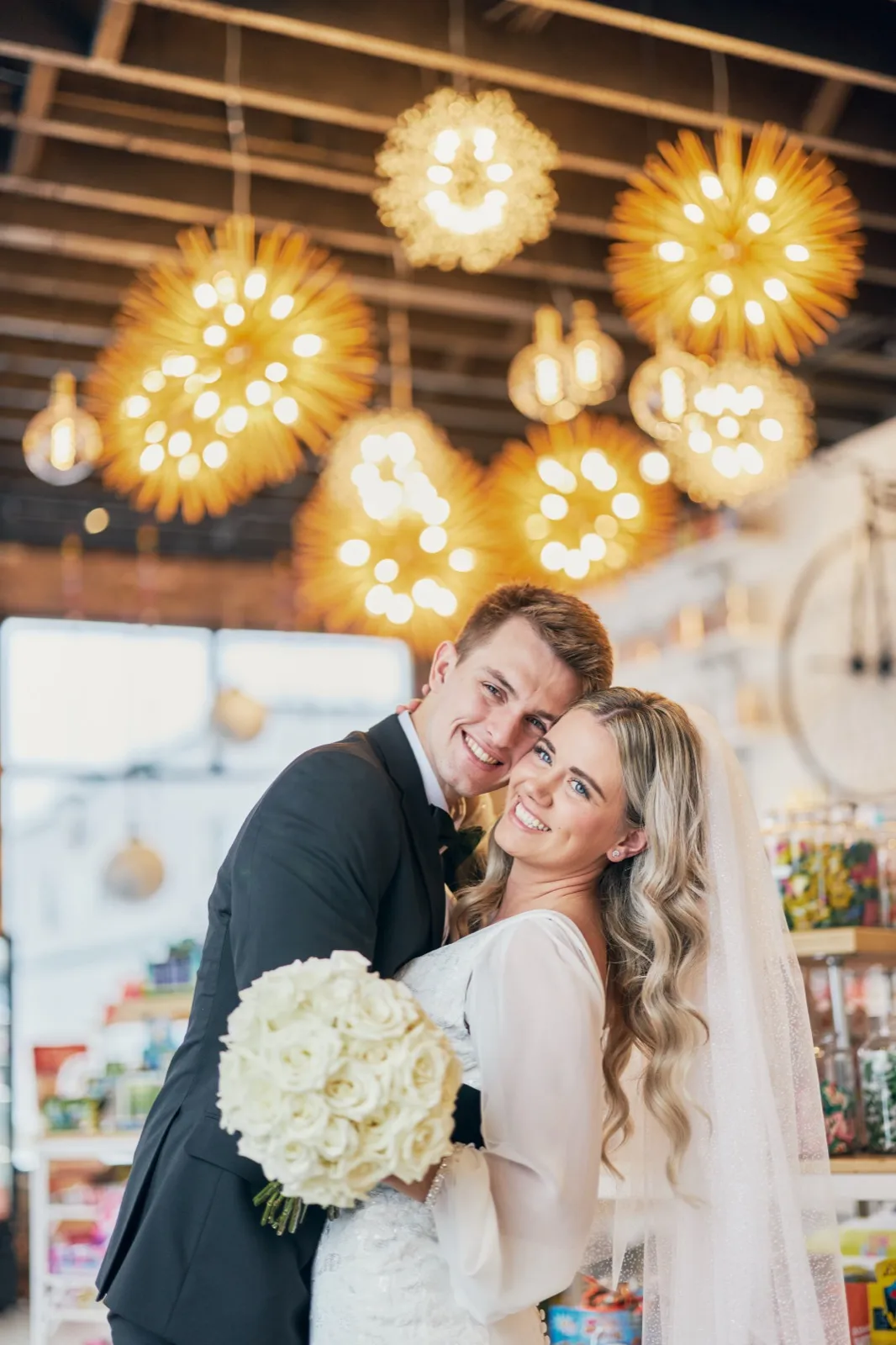 The bride and groom press cheek-to-cheek and smile at the camera beneath a cluster of starburst pendant lights, the bride holding a white rose bouquet. — Tim Larsen Photography, Brainerd Lakes MN