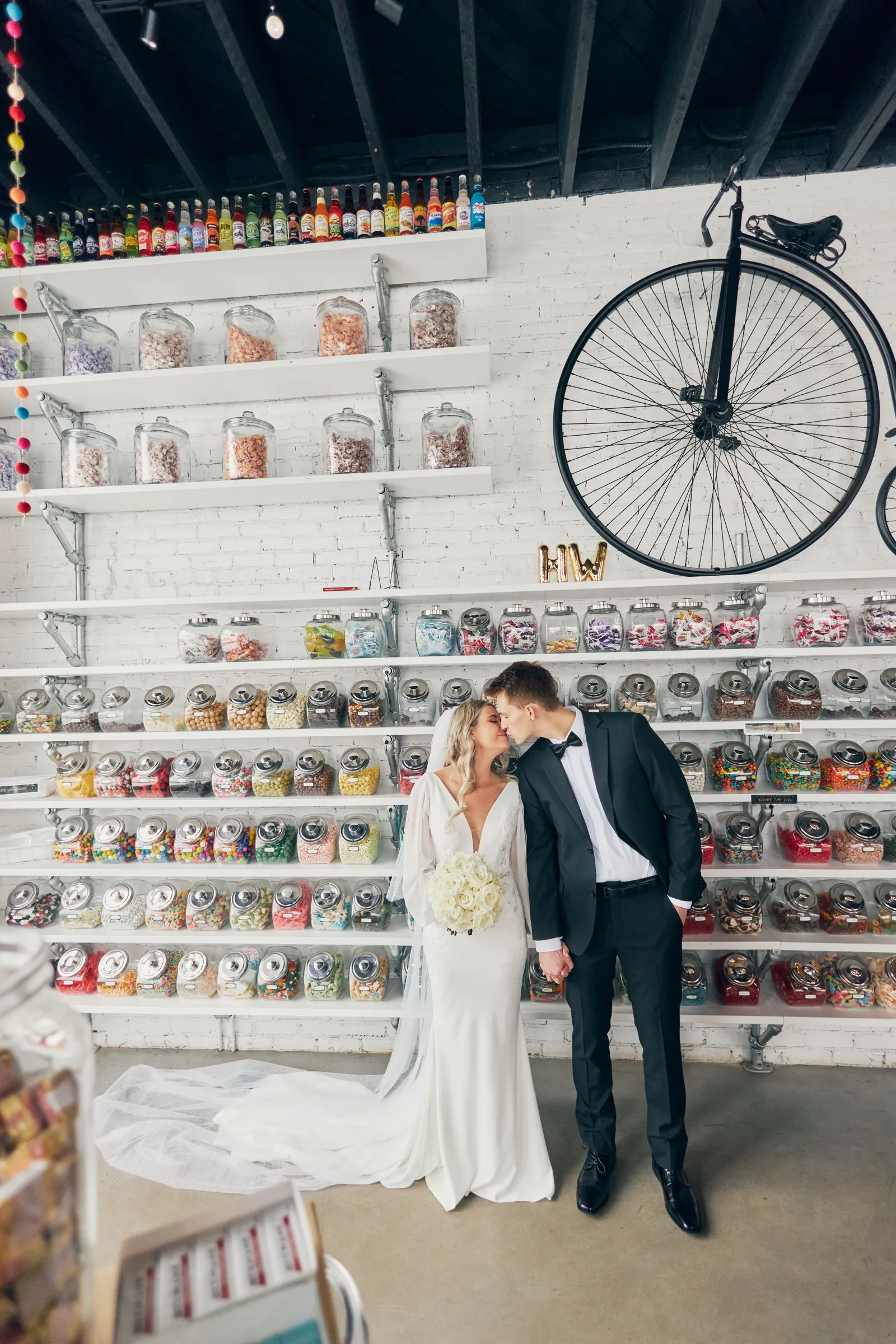 Madeline and Sam lean their foreheads together in a candy shop full of jars, a penny-farthing bicycle mounted on the white brick wall above them — Tim Larsen Photography, Brainerd Lakes MN