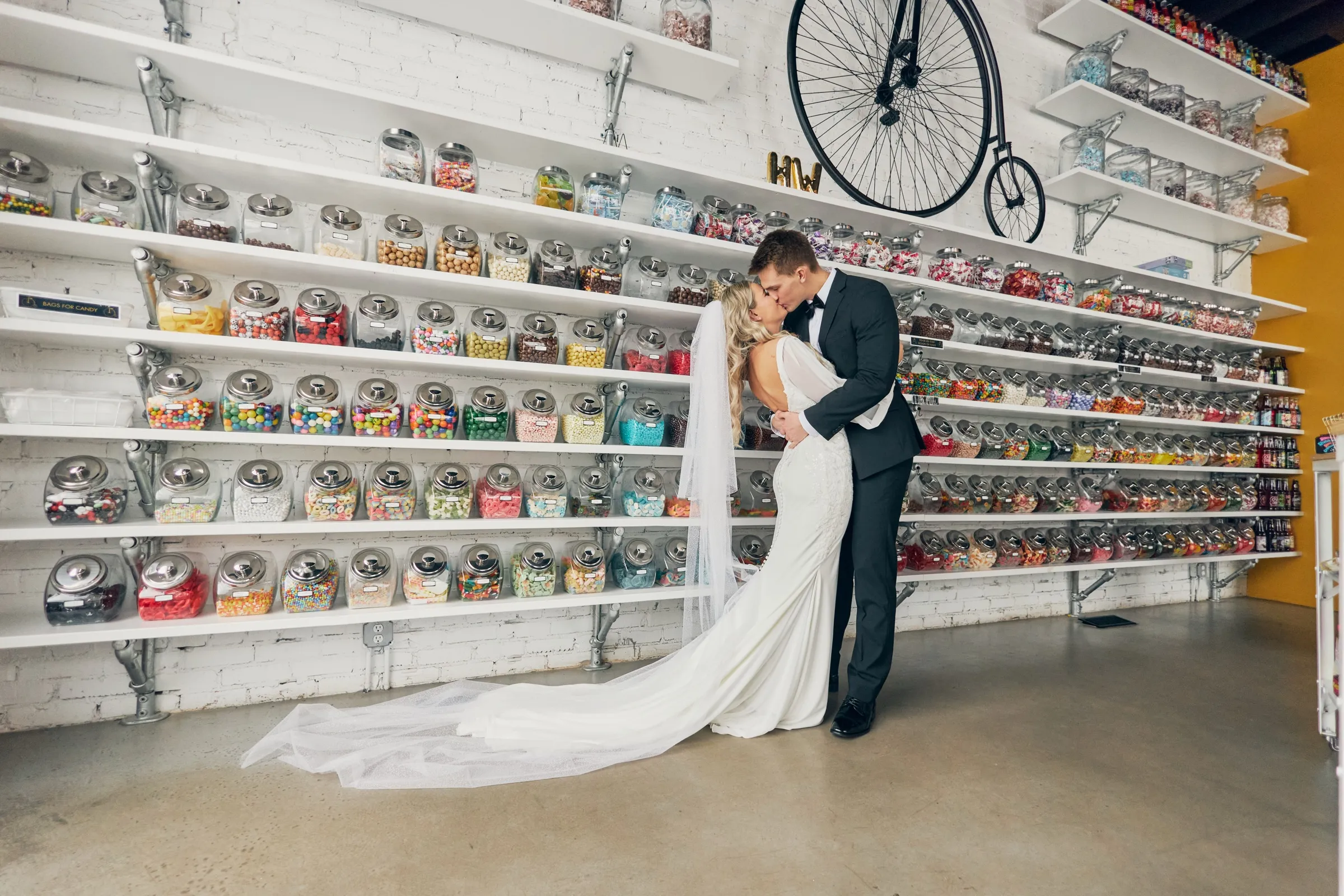 Madeline and Sam kiss in front of a brick wall lined with shelves of colorful candy jars and a vintage high-wheel bicycle — a downtown Crosby wedding portrait — Tim Larsen Photography, Brainerd Lakes MN