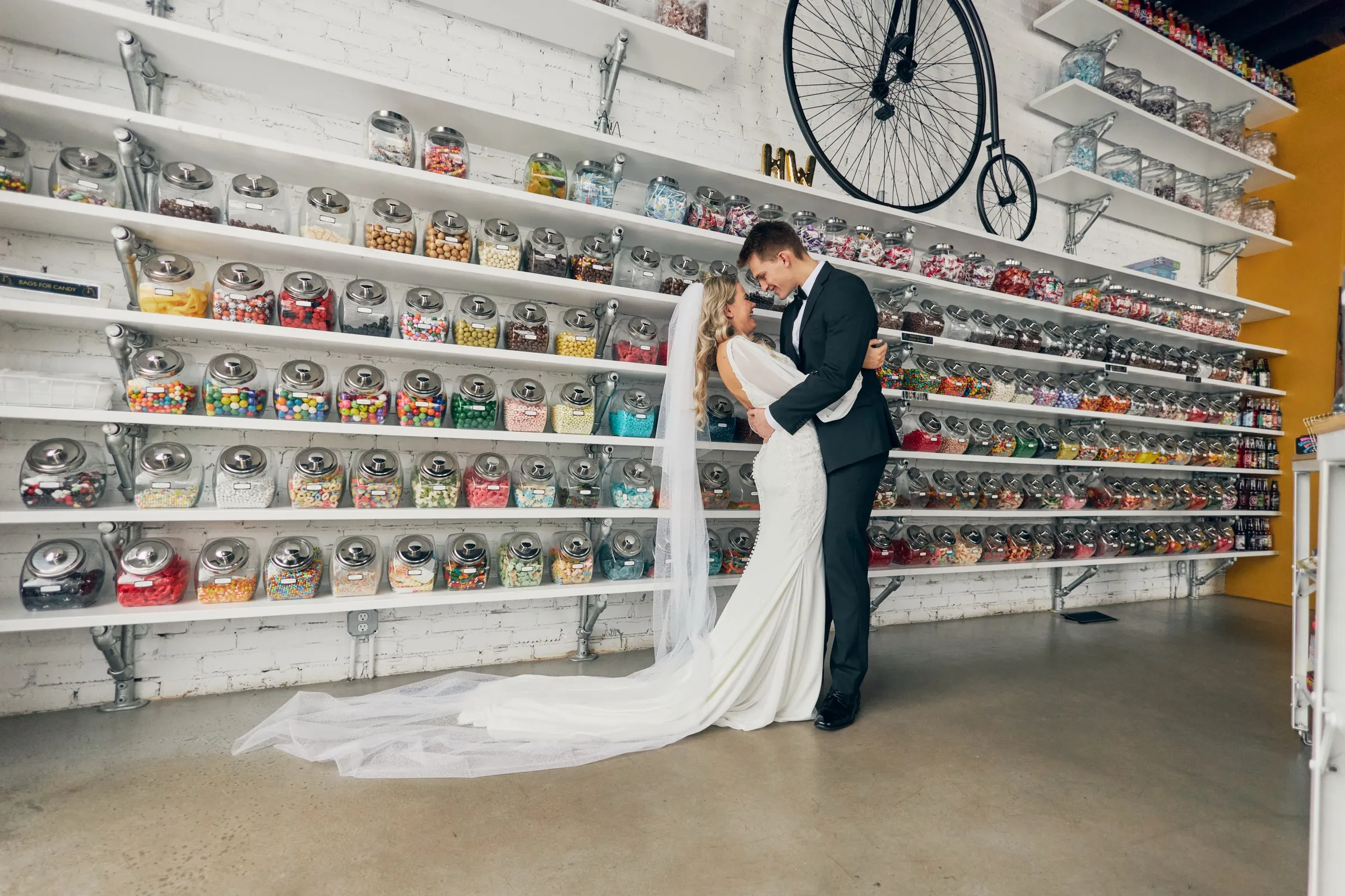 Madeline and Sam embrace forehead-to-forehead before a wall of colorful candy jars, her long veil and dress train trailing across the polished floor — Tim Larsen Photography, Brainerd Lakes MN