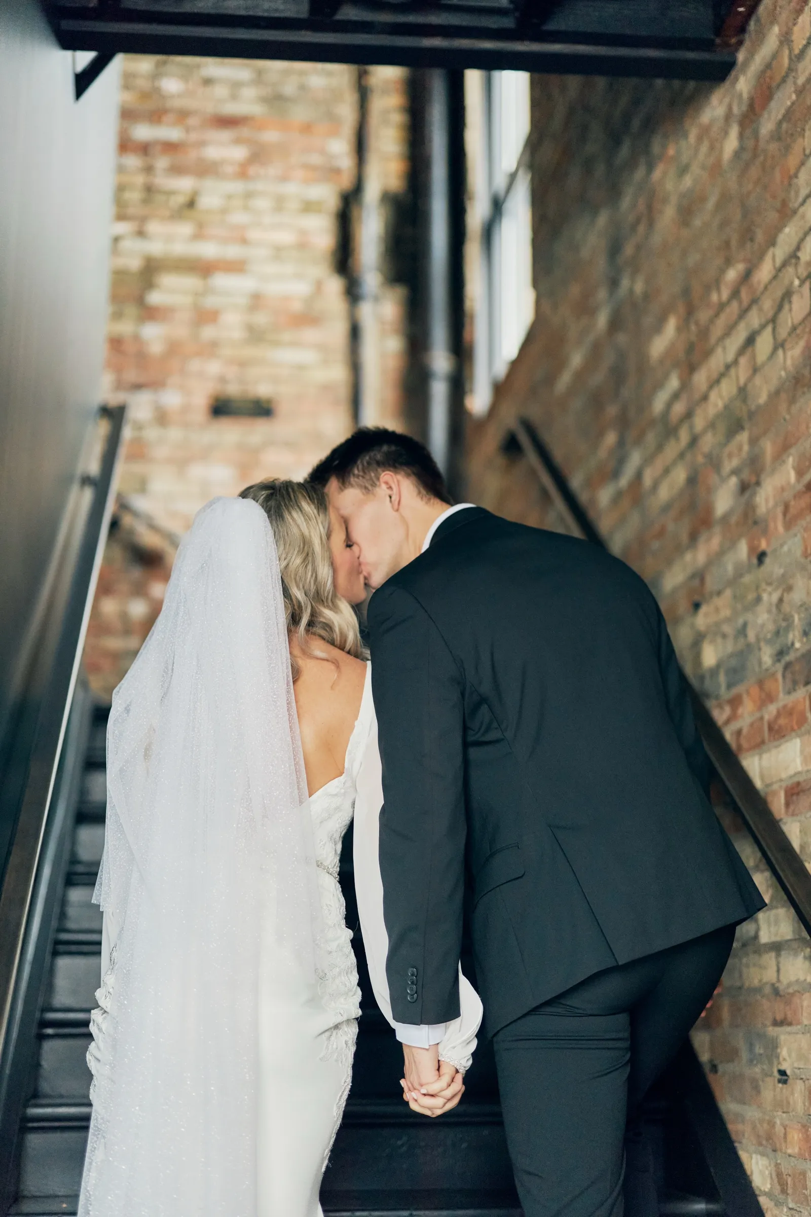 Madeline and Sam kiss on a brick-walled stairwell, her long veil trailing down the steps behind her — Tim Larsen Photography, Brainerd Lakes MN
