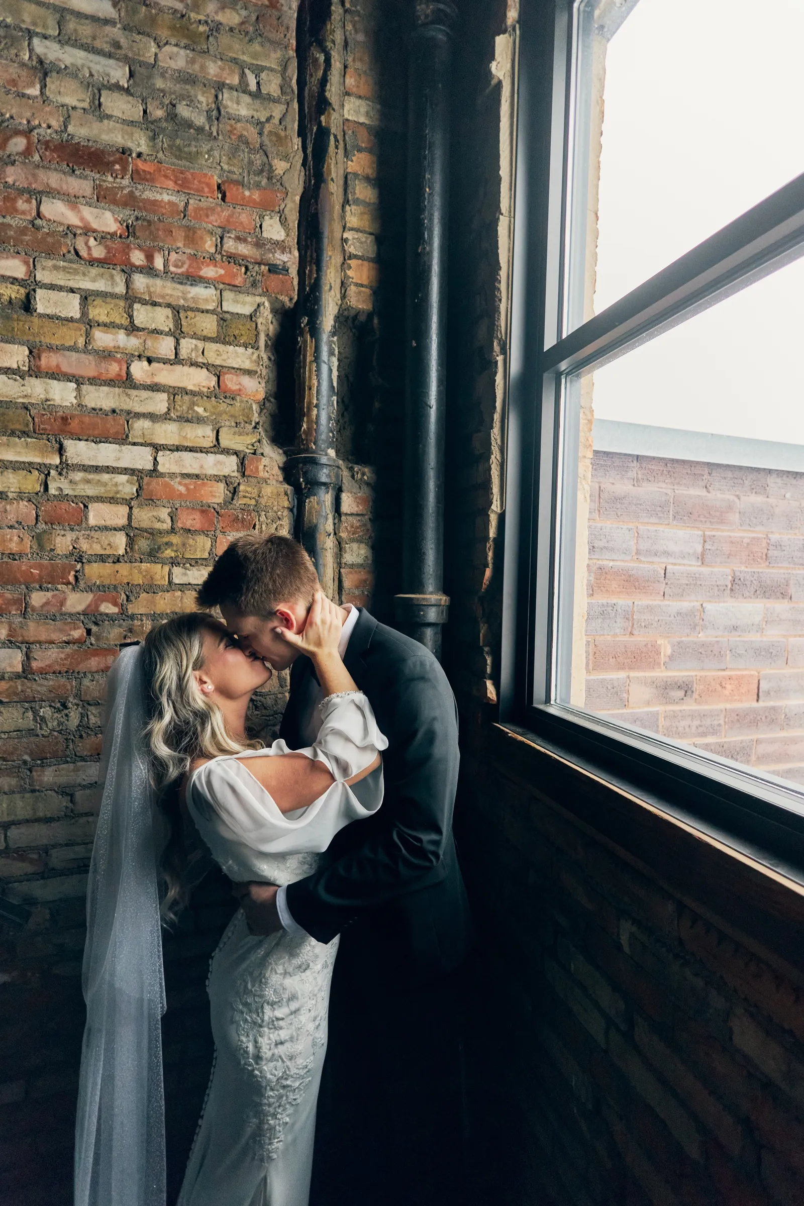Madeline and Sam kiss beside a tall industrial window, framed by a weathered brick wall and exposed pipe — Tim Larsen Photography, Brainerd Lakes MN