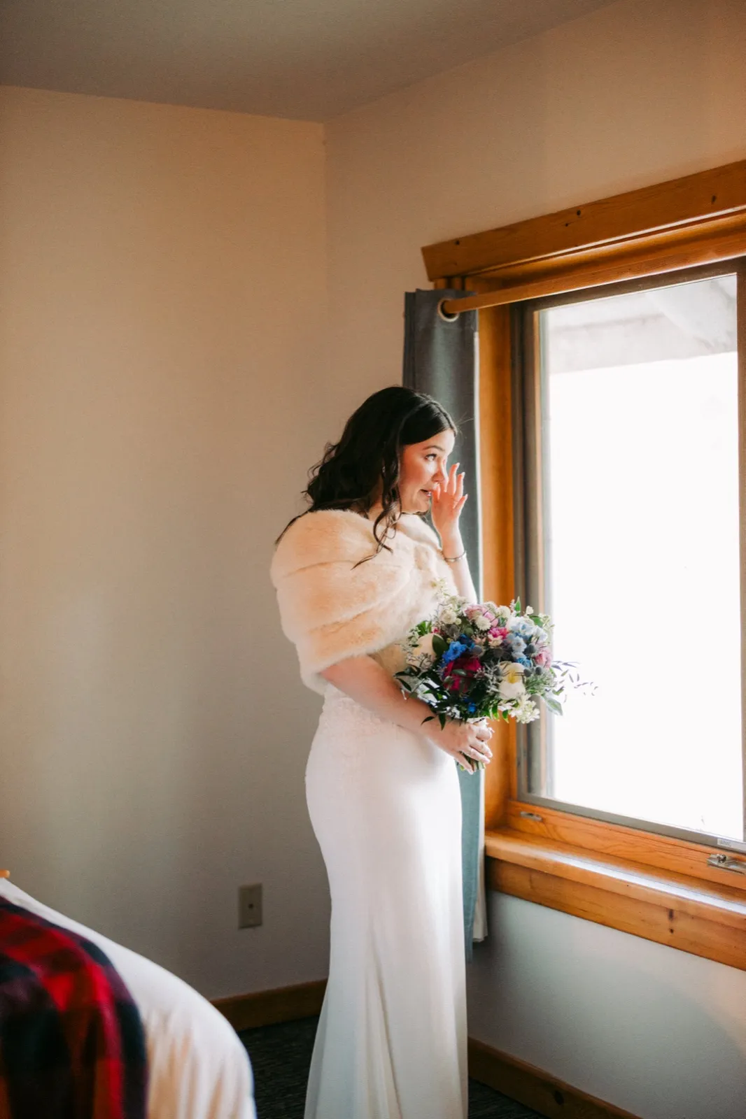 The bride in a white gown and fur stole holds her bouquet and wipes a tear while standing in window light in the bridal suite — Tim Larsen Photography, Brainerd Lakes MN