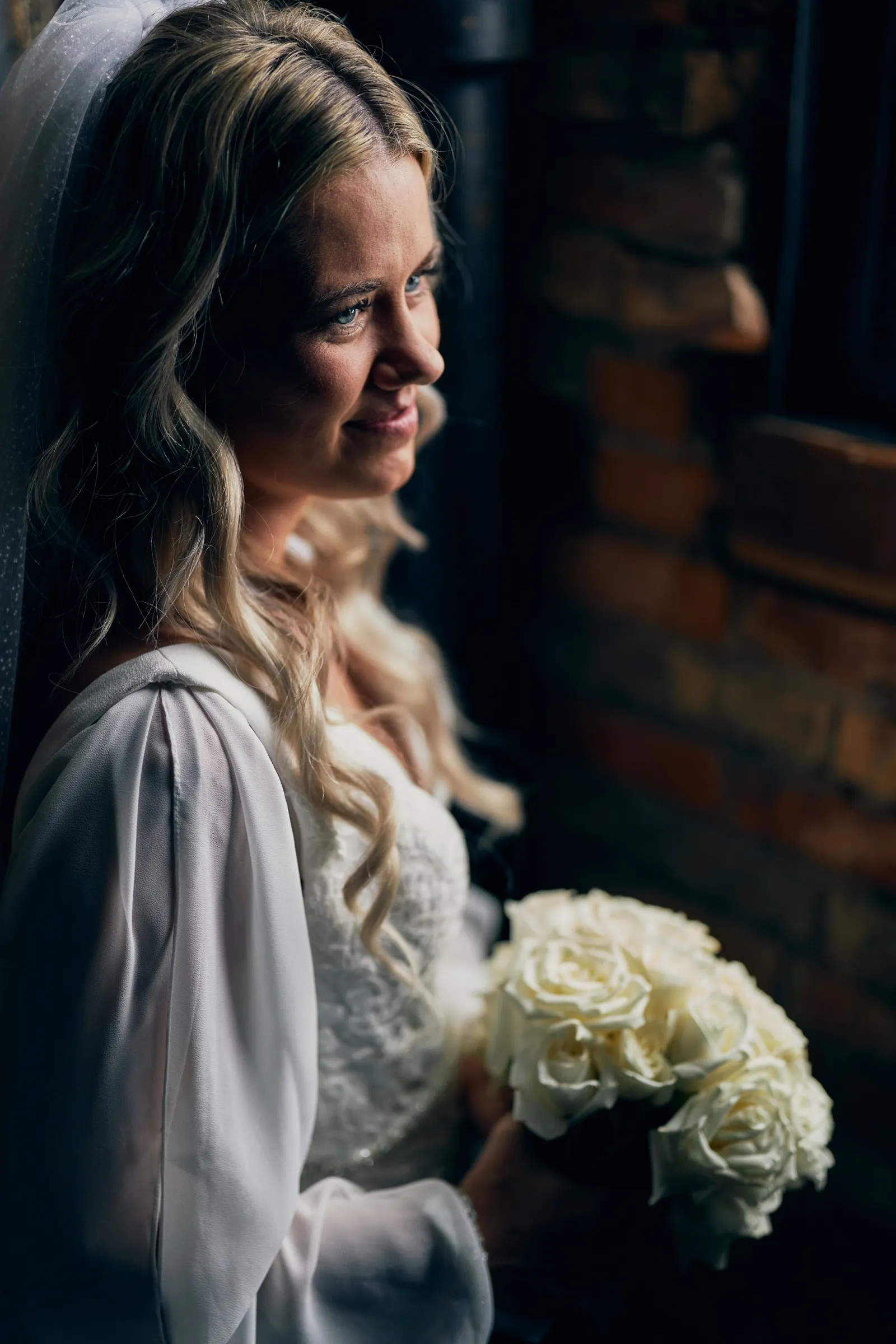 Madeline stands beside a window in soft side-light, holding a white rose bouquet, her veil draped over her shoulder against an exposed brick wall — Tim Larsen Photography, Brainerd Lakes MN