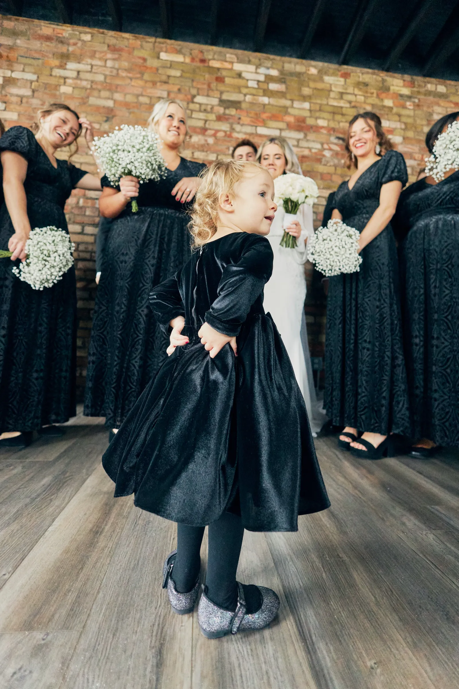 A young flower girl in a black velvet dress poses with her hands on her hips while Madeline and the bridesmaids laugh behind her in a brick-walled room — Tim Larsen Photography, Brainerd Lakes MN