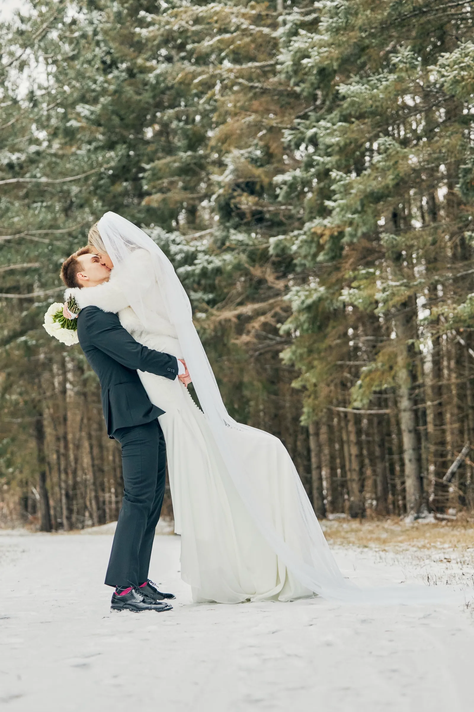 Sam lifts Madeline into a kiss in a snowy clearing, her long veil and dress trailing across the snow with pine trees behind them — Tim Larsen Photography, Brainerd Lakes MN