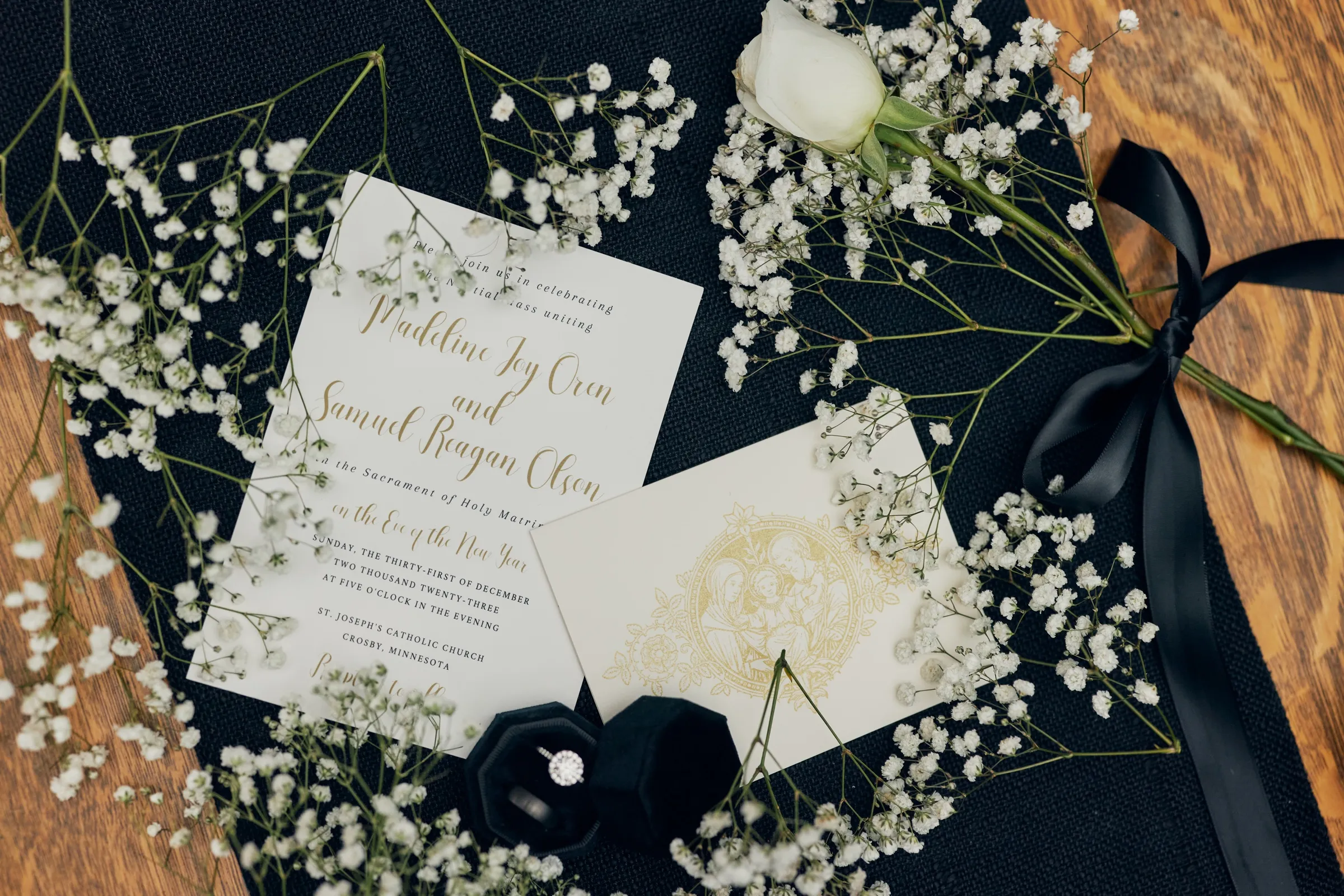 A flat-lay of the wedding invitation, gold-foil envelope, a white rose boutonniere, baby's breath, black ribbon, and Madeline's diamond ring in a dark velvet box — Tim Larsen Photography, Brainerd Lakes MN